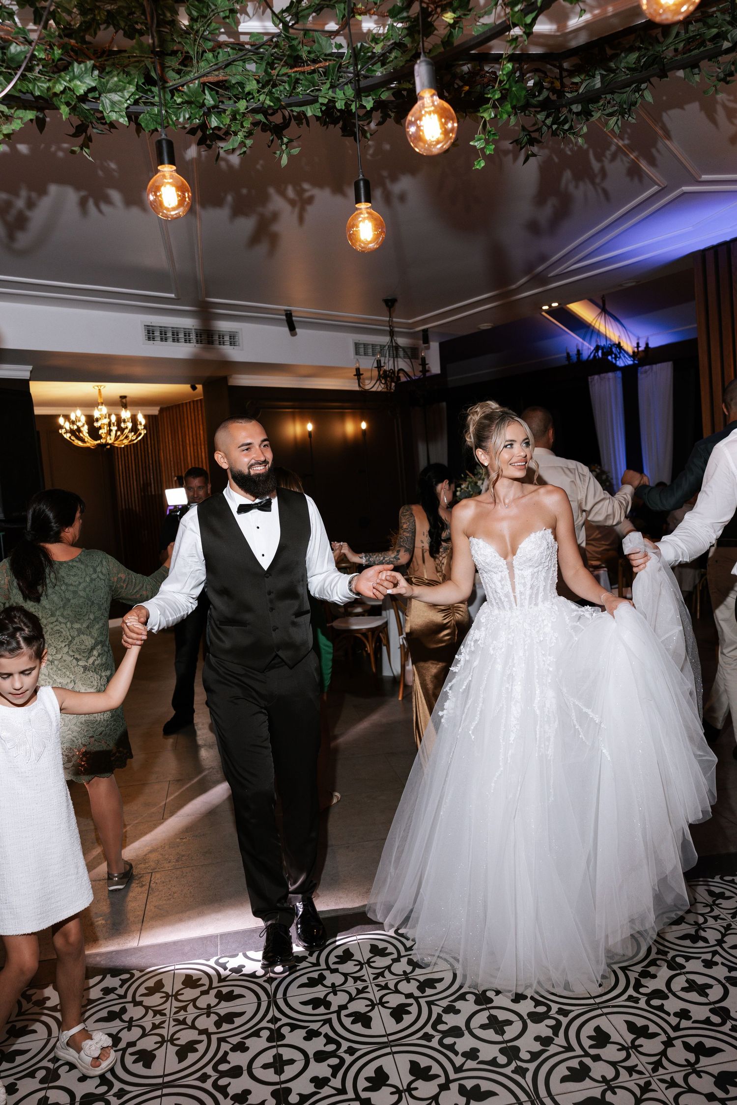 Wedding guests dancing and celebrating on a black and white patterned dance floor under hanging Edison bulbs and greenery.