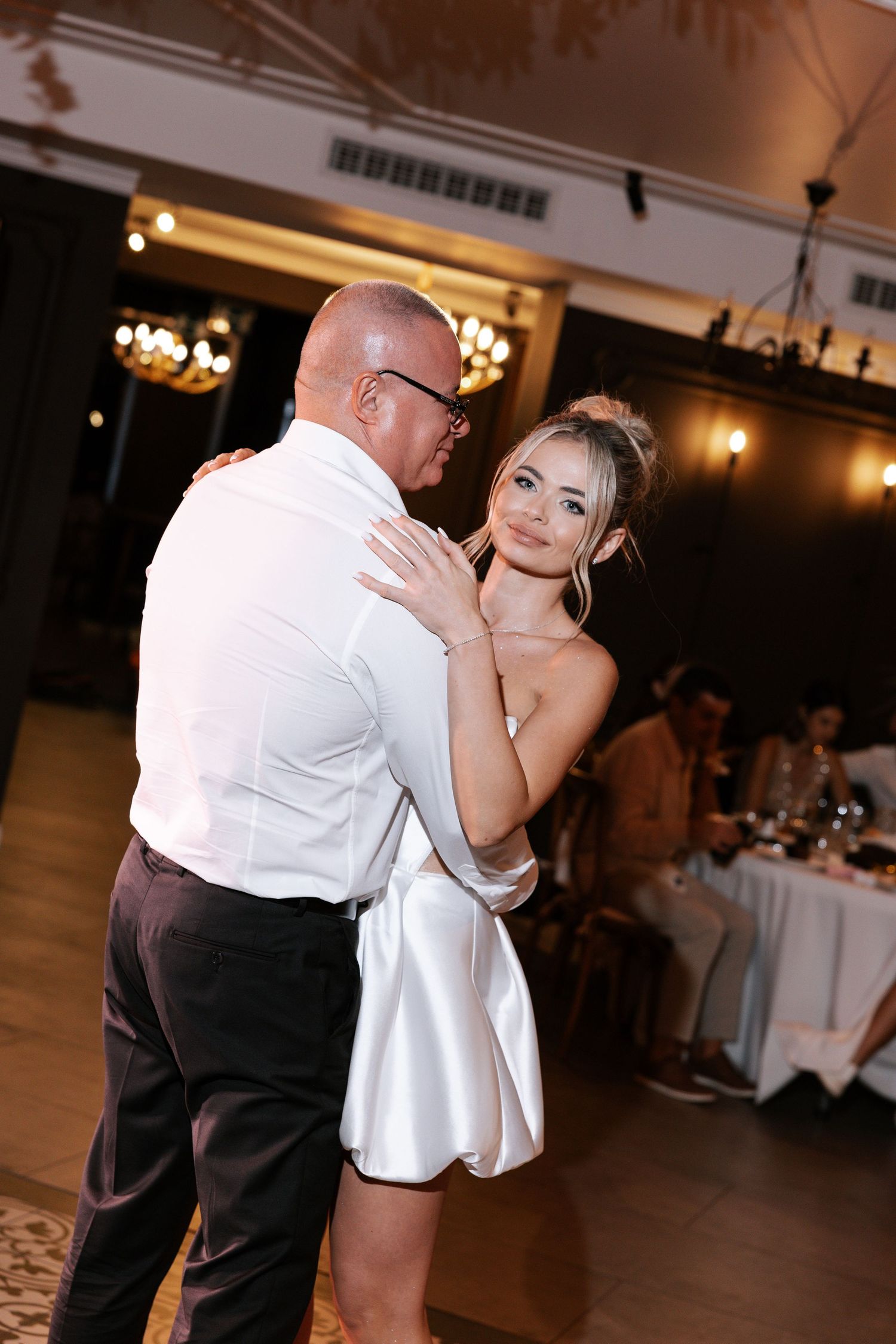 A couple shares an intimate dance moment in a dimly lit reception hall with elegant chandeliers and white tablecloths.