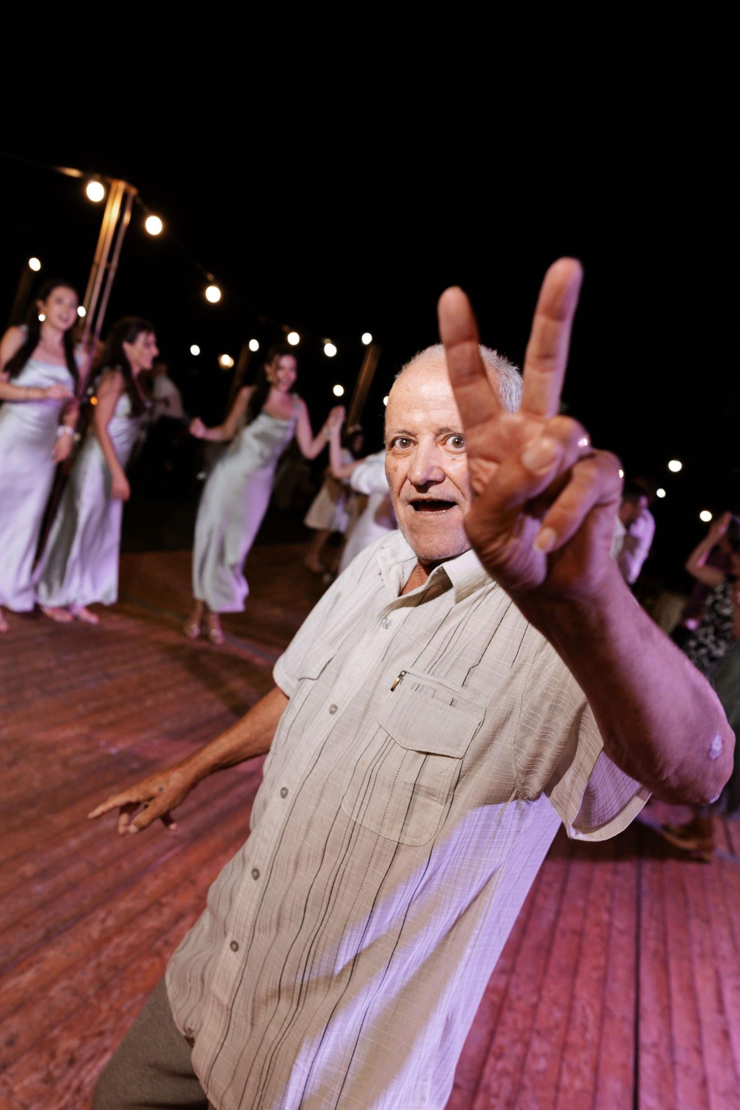 People dancing at a wedding reception on a rustic wooden dance floor illuminated by string lights above.