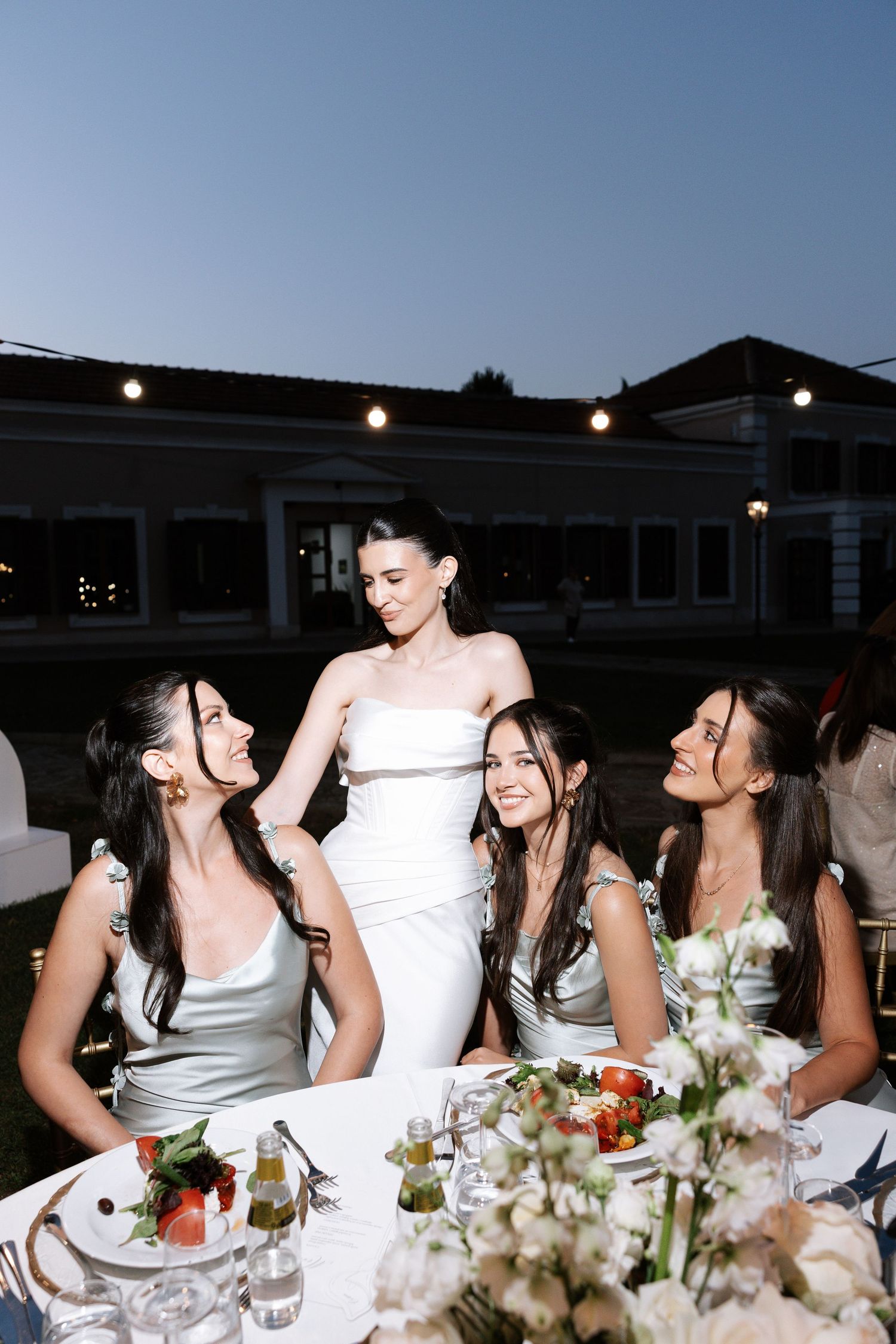 A group of bridesmaids in light colored dresses share laughter at an elegant wedding reception table.