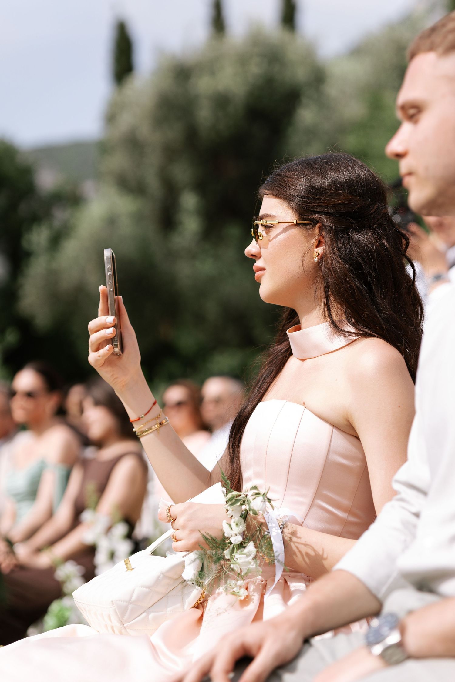 Wedding guests in pink and white attire sit outdoors while taking photos on their phones during a ceremony.