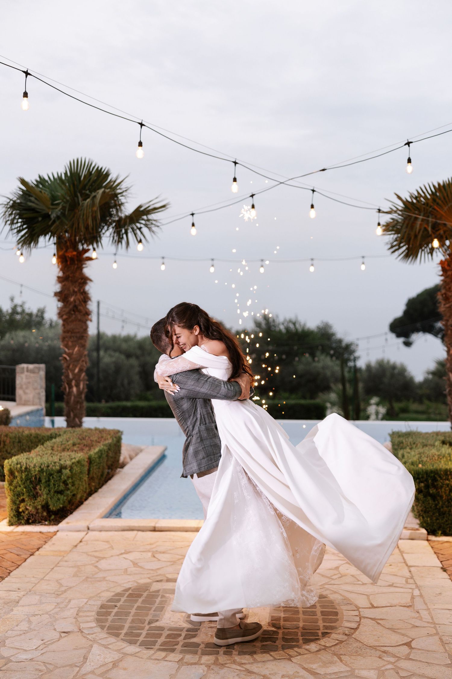 A couple performs a romantic dip dance move on a stone patio under twinkling string lights at sunset.