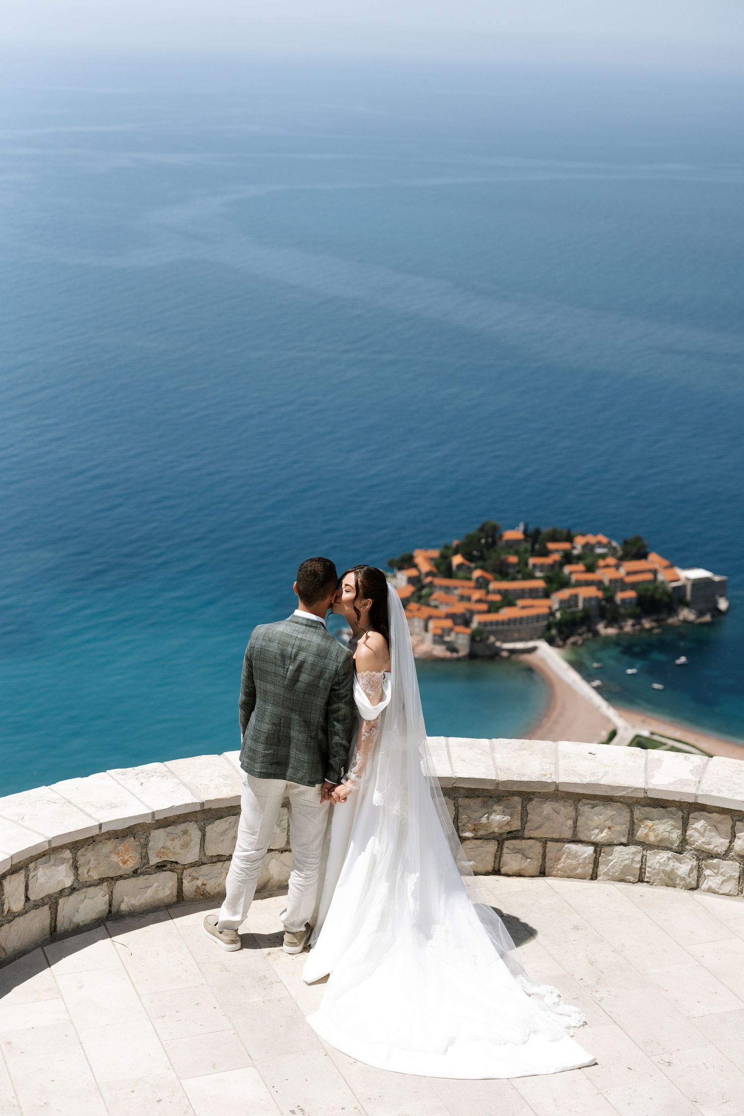 A couple in wedding attire stands at a scenic overlook with views of Sveti Stefan island resort in Montenegro.