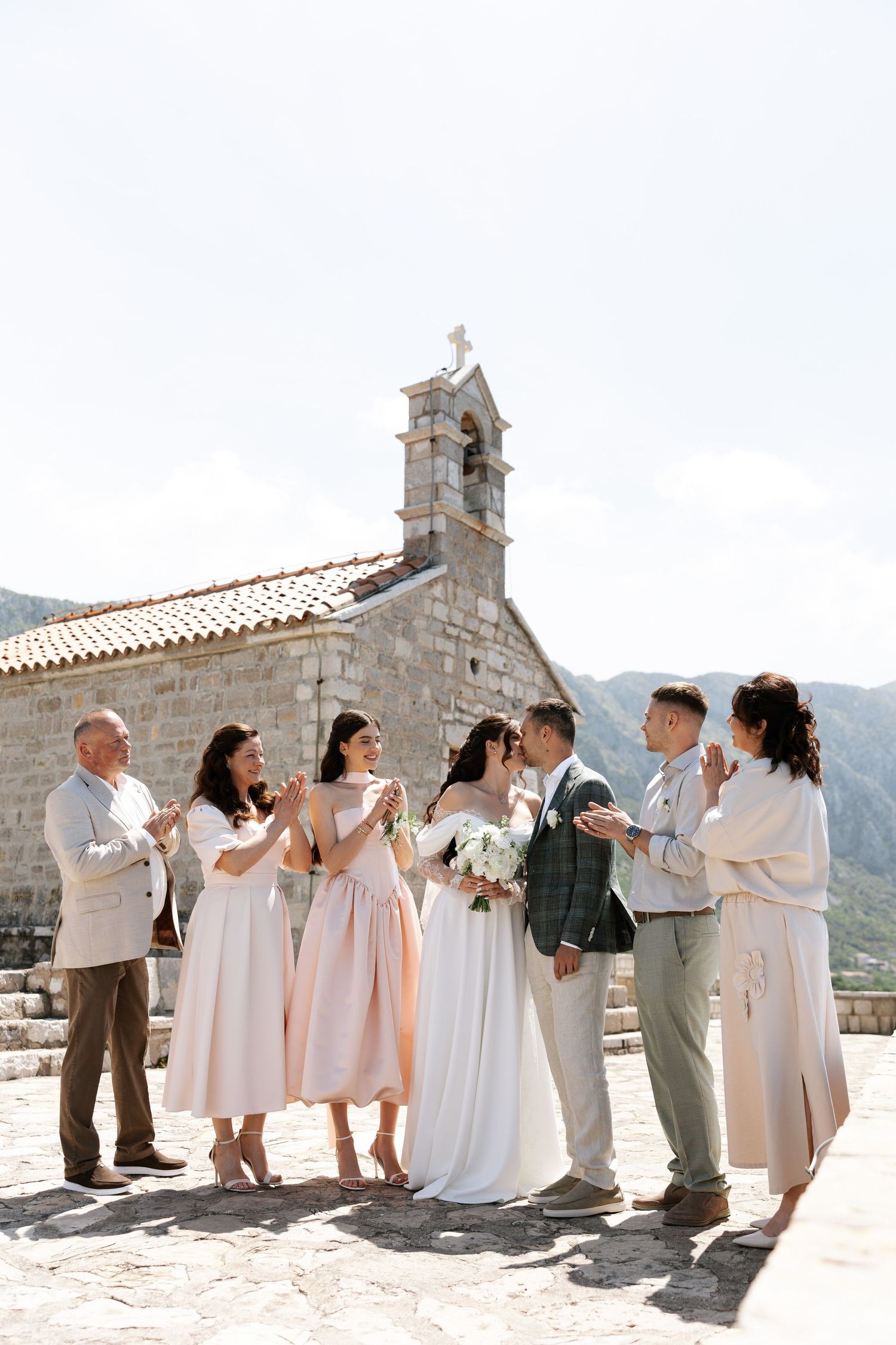 Wedding party celebrates together outside a rustic stone church with mountain backdrop in Montenegro.
