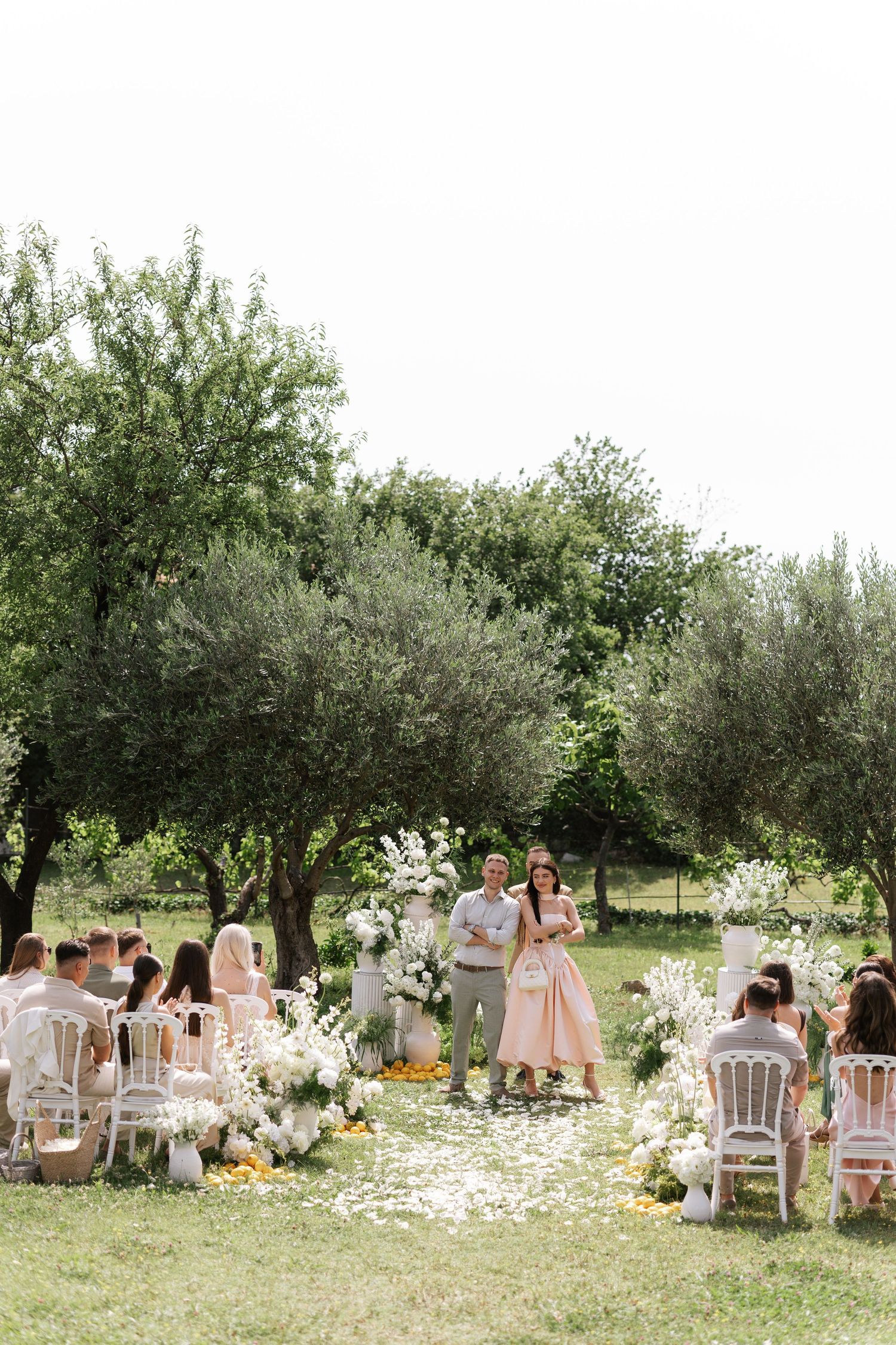 An outdoor wedding ceremony setup in an olive grove with white chairs and floral decorations scattered on the grass.