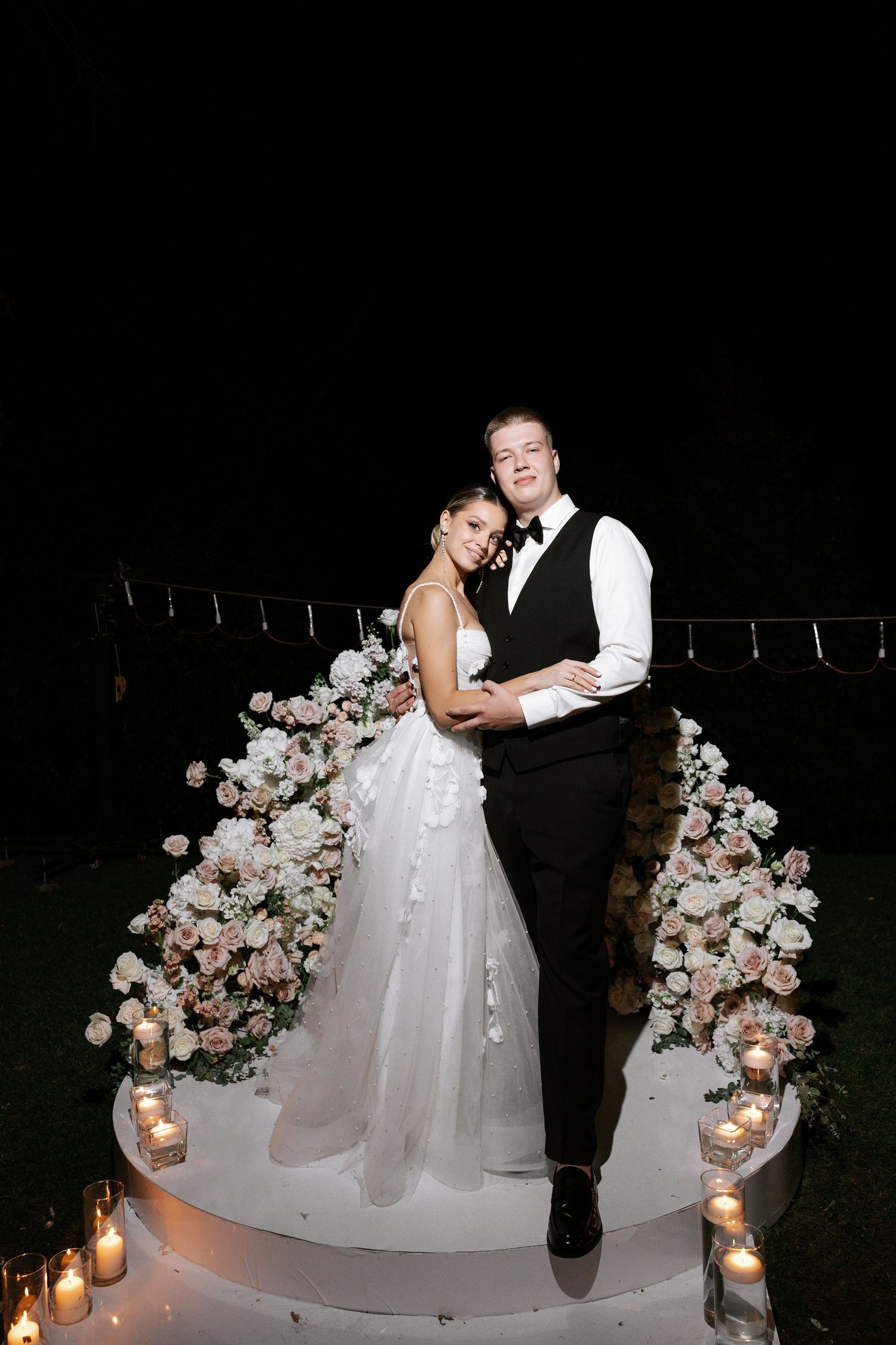 Romantic nighttime wedding portrait under floral arch with candle lighting.