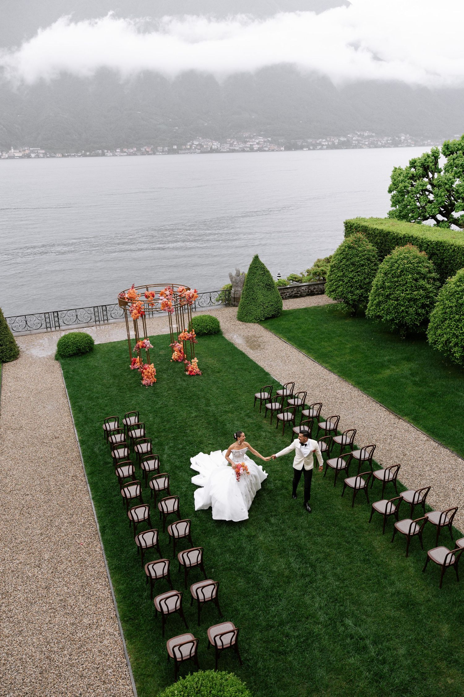 Outdoor wedding ceremony setup with rows of chairs on a green lawn overlooking a misty lake and mountains in the background.