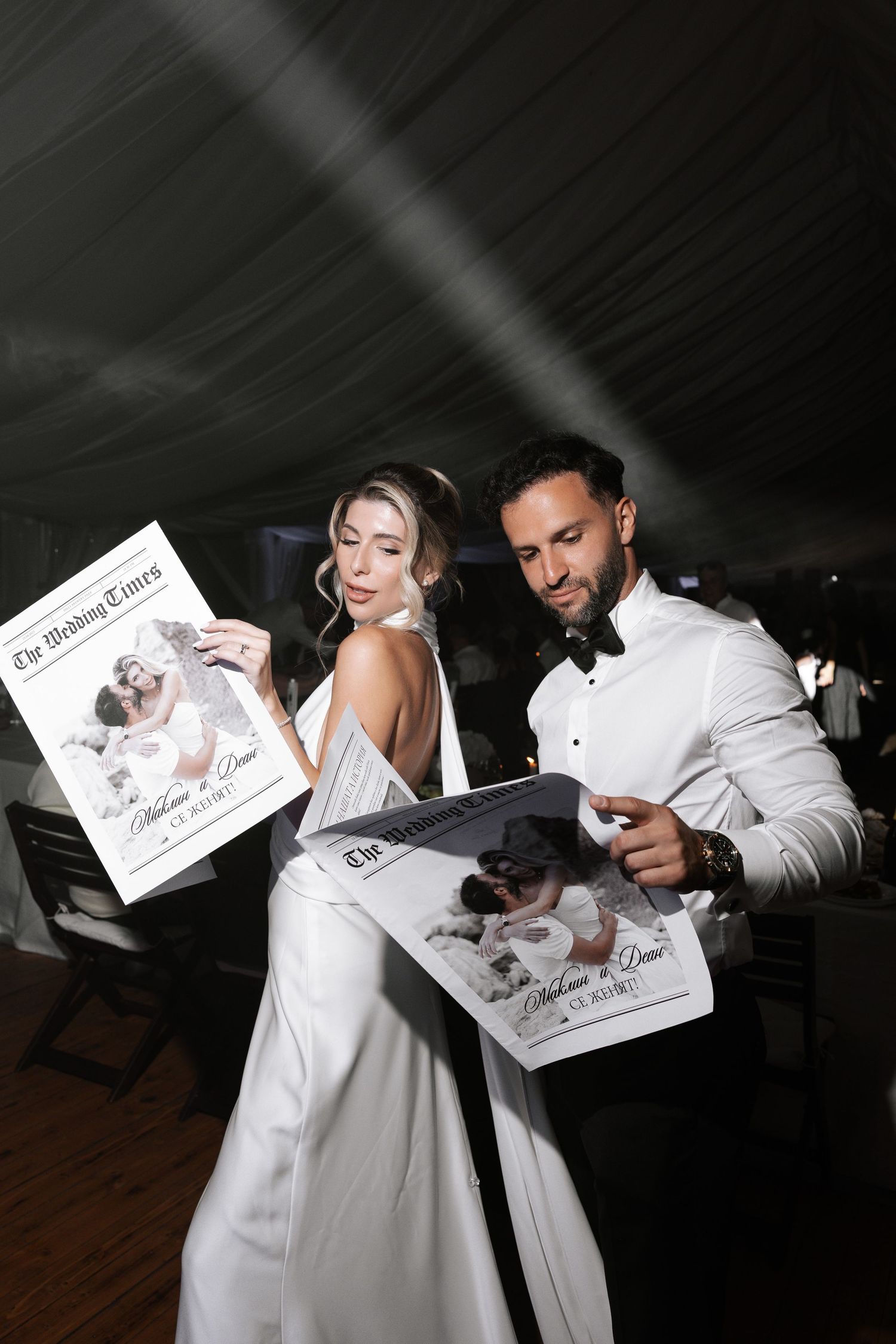 Two people in white outfits share a playful moment while looking at paper documents in a dimly lit setting.