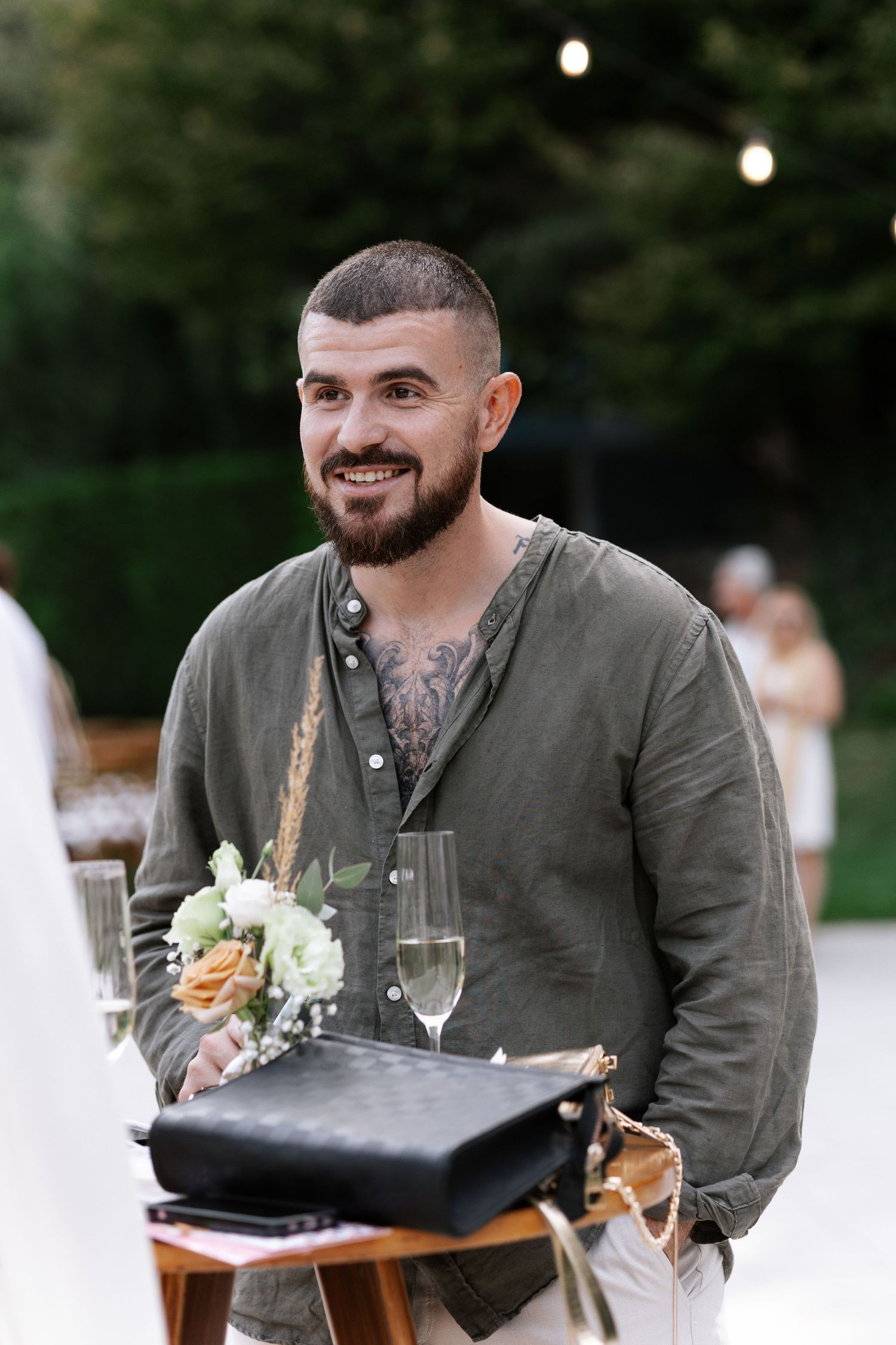A smiling person in an olive green henley shirt holds flowers and a black folder at an outdoor event.