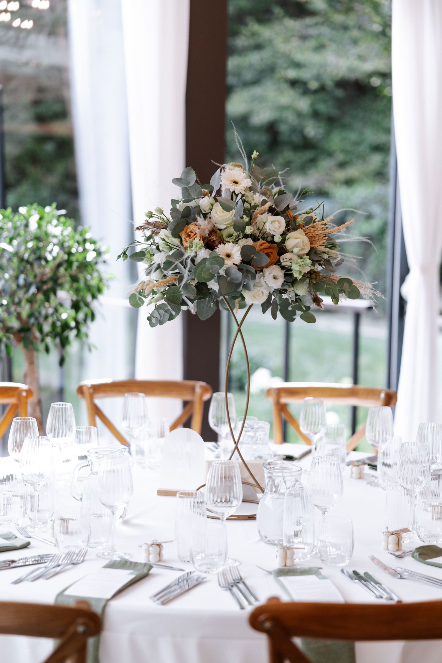 Elegant wedding reception table setting with tall floral centerpiece featuring eucalyptus and roses in a glass vase.