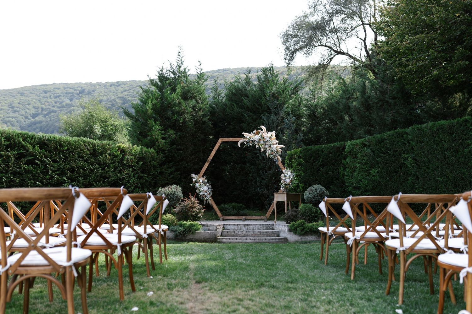 Rustic wooden cross-back chairs line up in rows on a grassy hillside for an outdoor wedding ceremony.