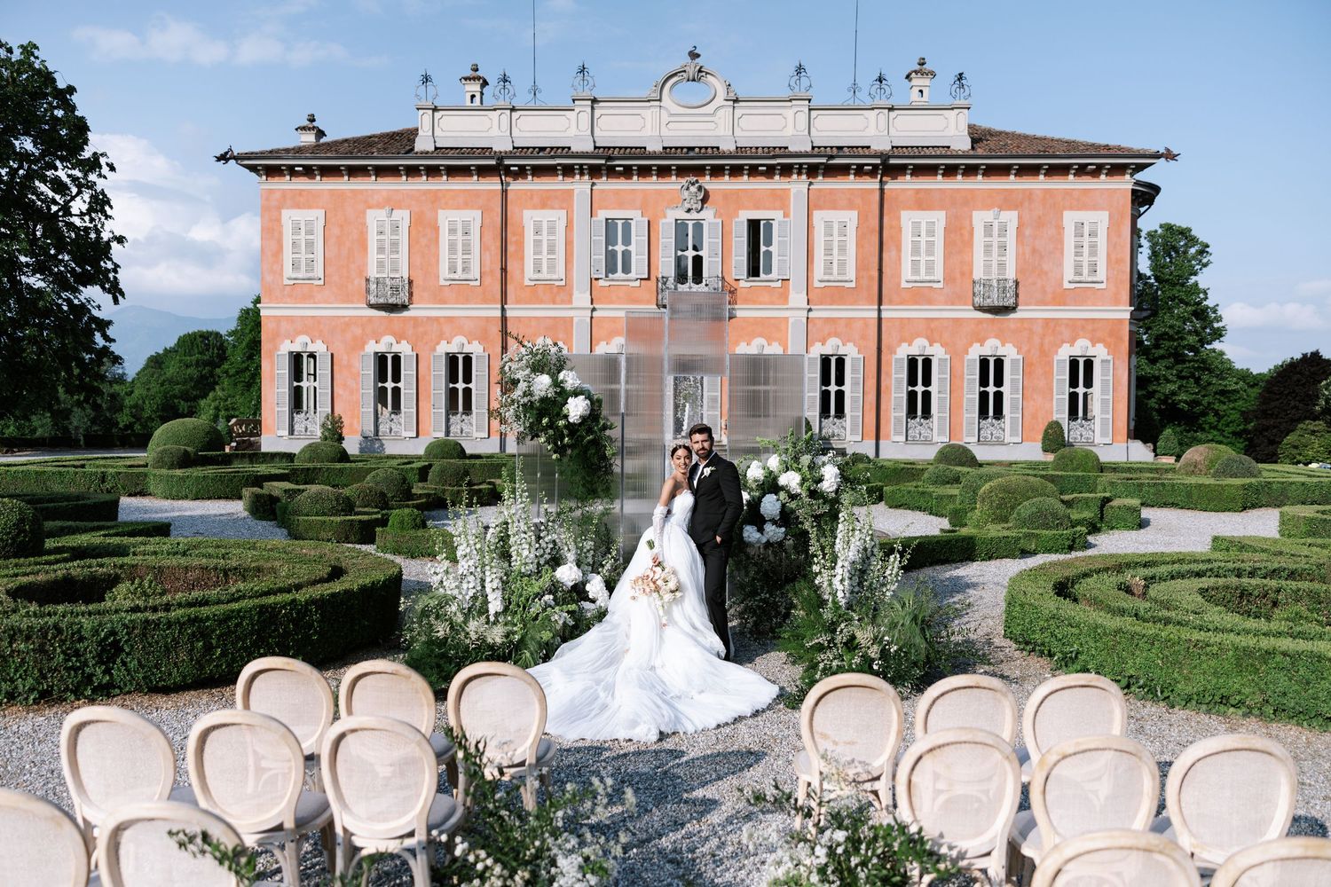 Elegant outdoor wedding ceremony setup with white chairs and pink mansion backdrop in a manicured garden setting.