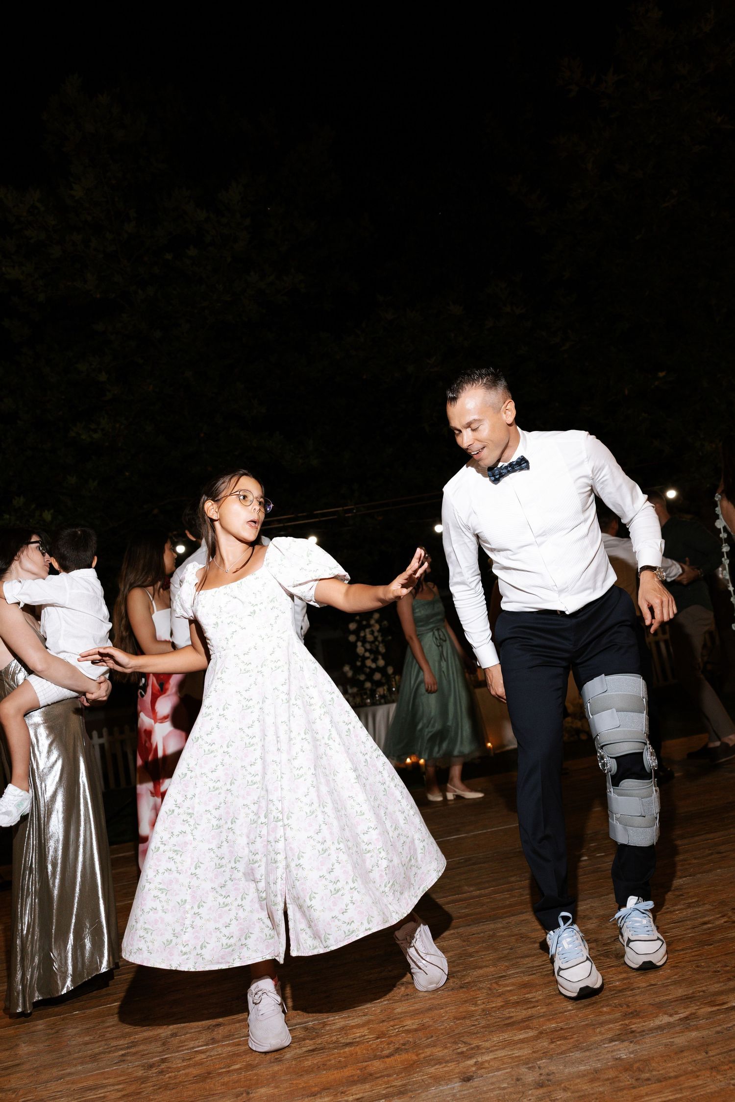 A couple dances energetically on a wooden dance floor, with the woman wearing a white dress and both in white sneakers.