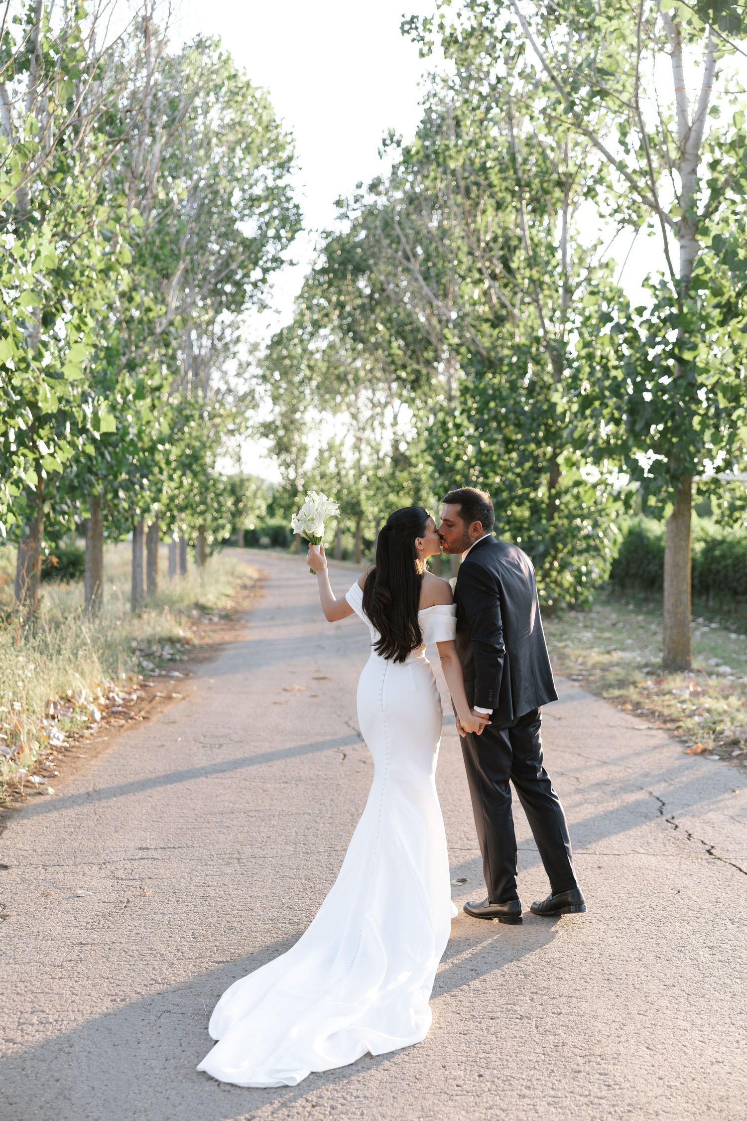 A couple shares a romantic moment on a tree-lined path, with the bride wearing a flowing white mermaid gown.
