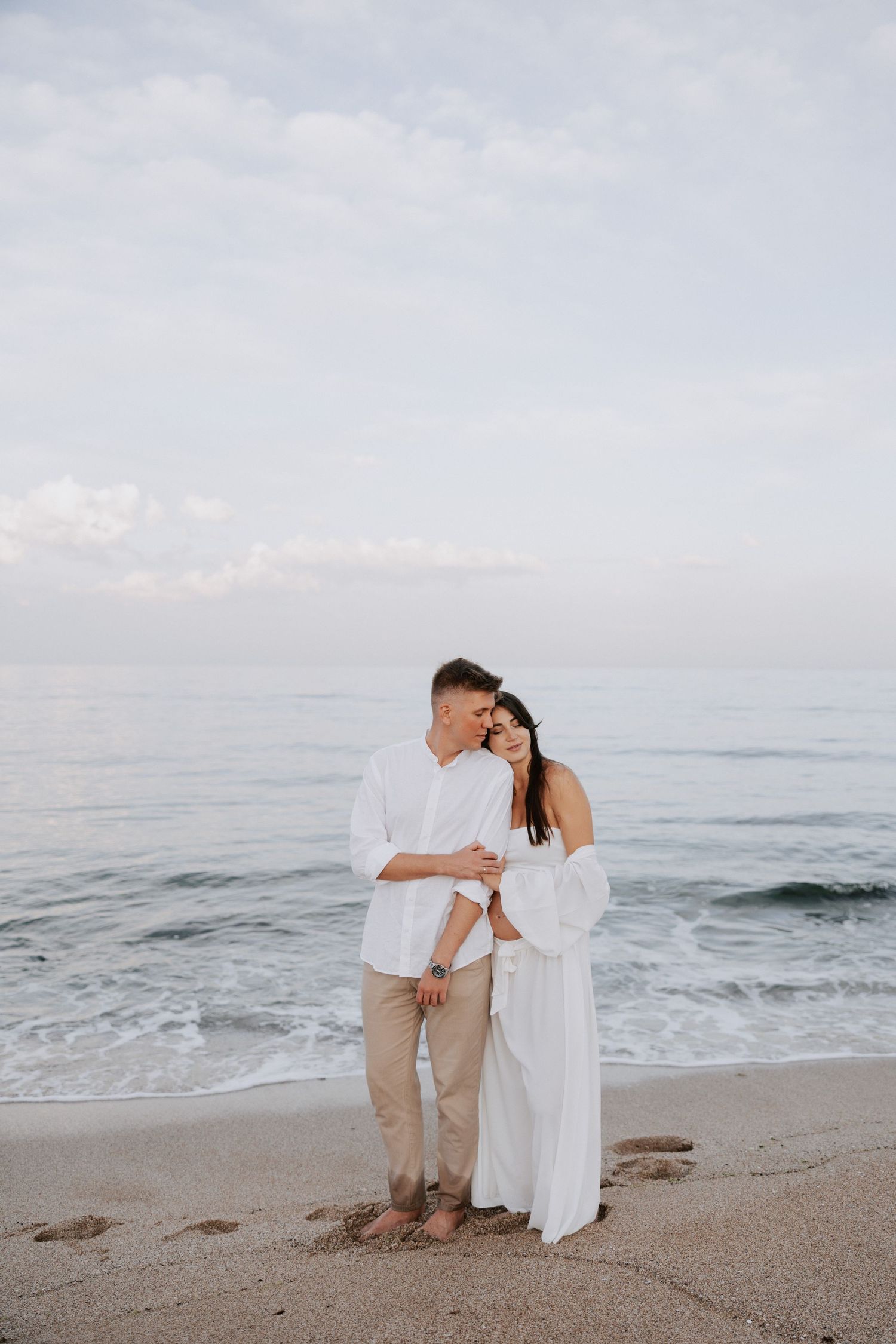 A couple embraces on a sandy beach against the ocean backdrop in coordinated white outfits.