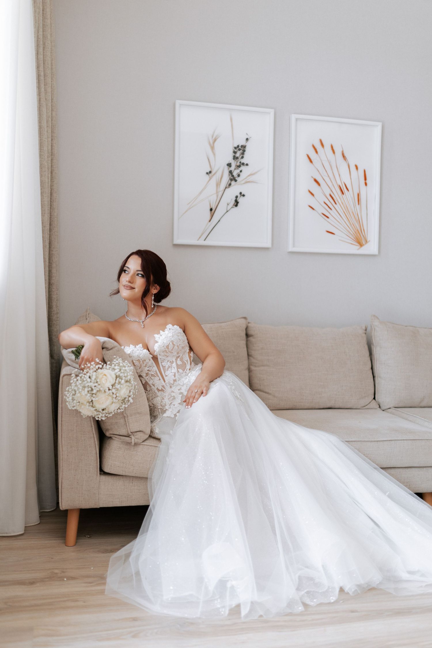 Elegant bride in a white strapless gown reclines on a beige couch with minimalist artwork in the background.