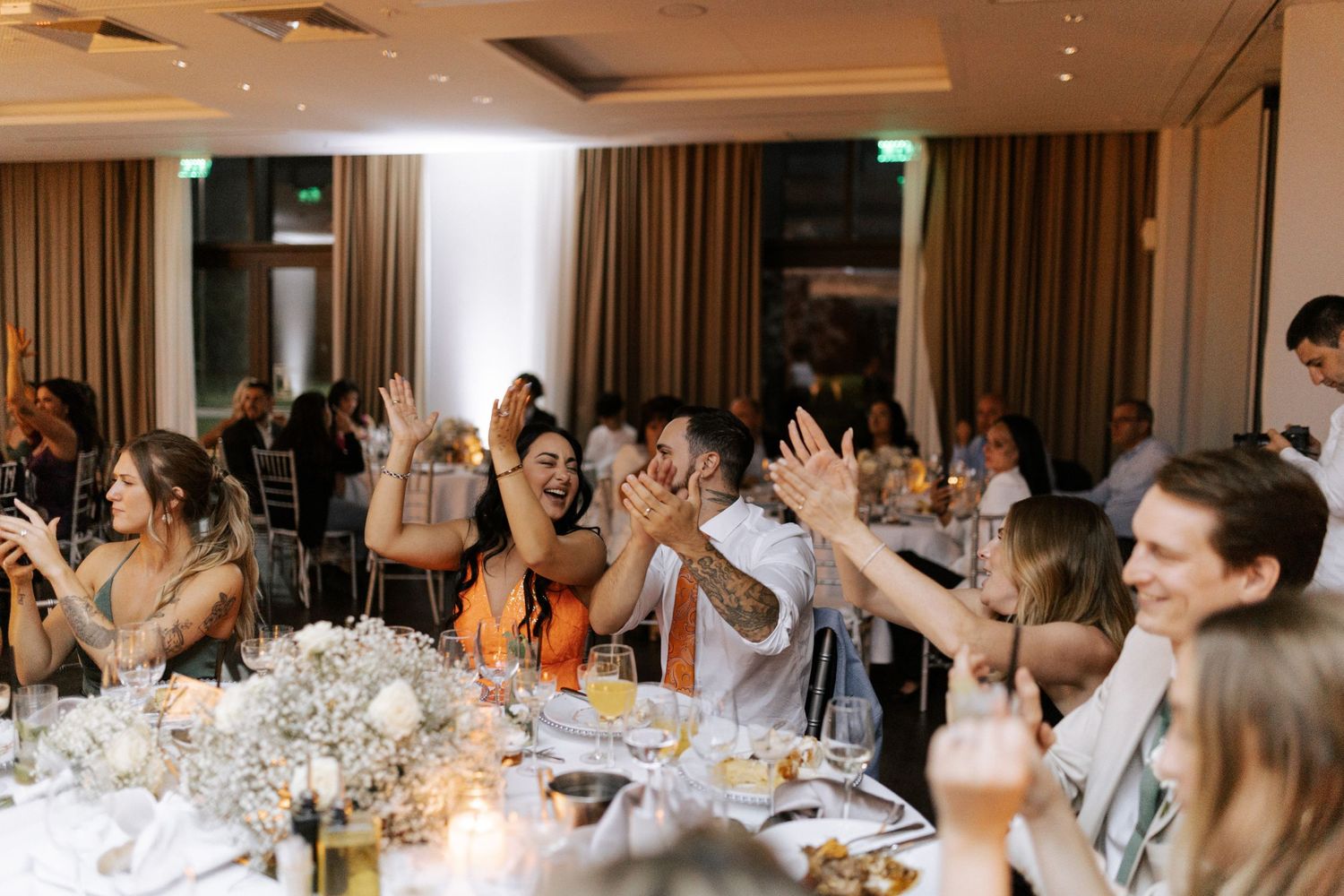 Wedding reception guests raise their hands in celebration at elegant dining tables with floral centerpieces.