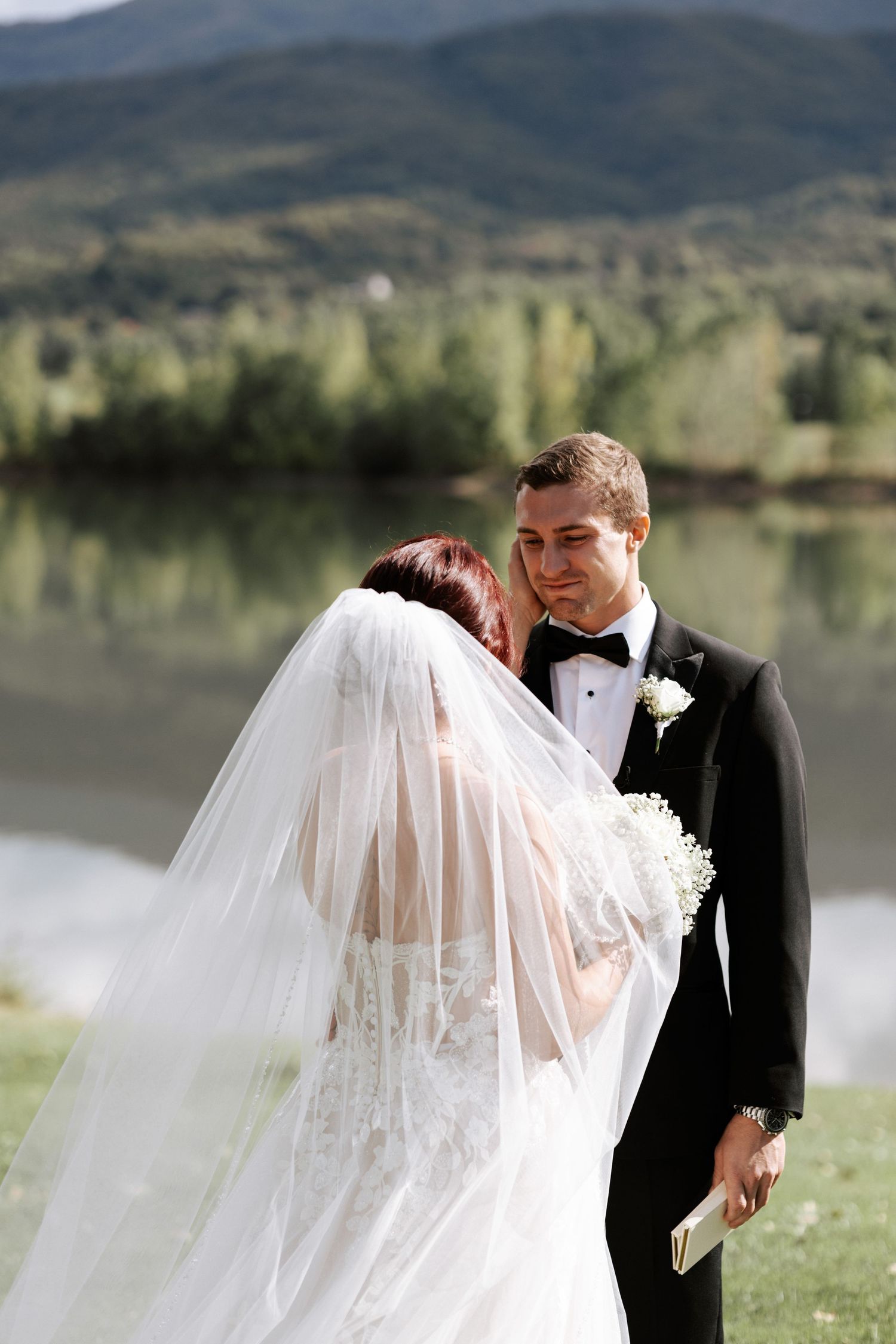 A romantic wedding couple shares an intimate moment by a serene lake with mountains in the background.
