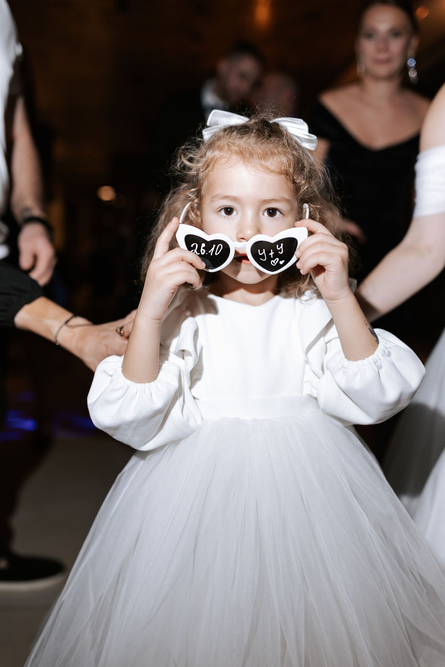 A young flower girl in white tulle dress playfully poses with heart-shaped sunglasses.