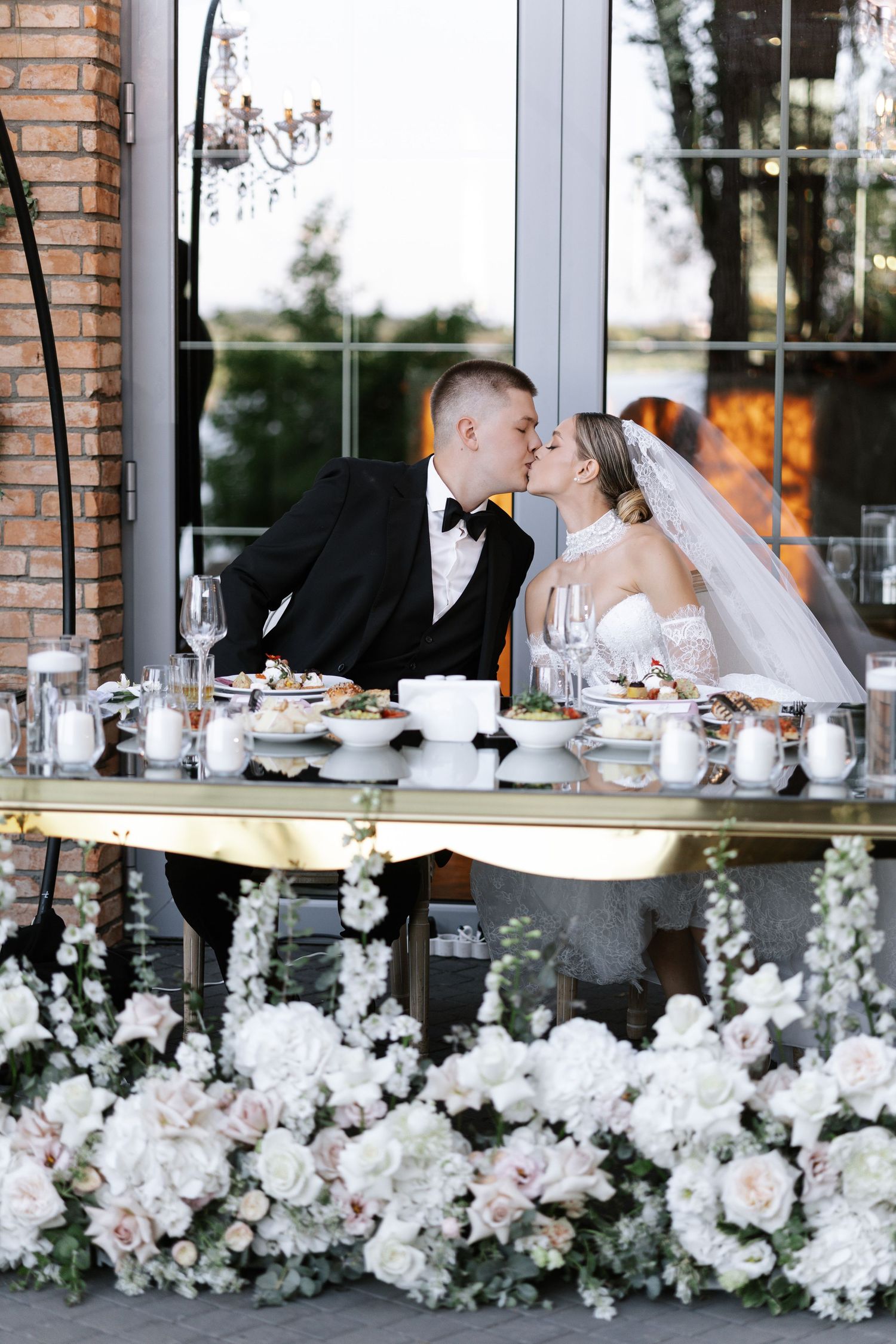 Newlyweds share an intimate moment at an elegantly decorated reception table with floral arrangements.