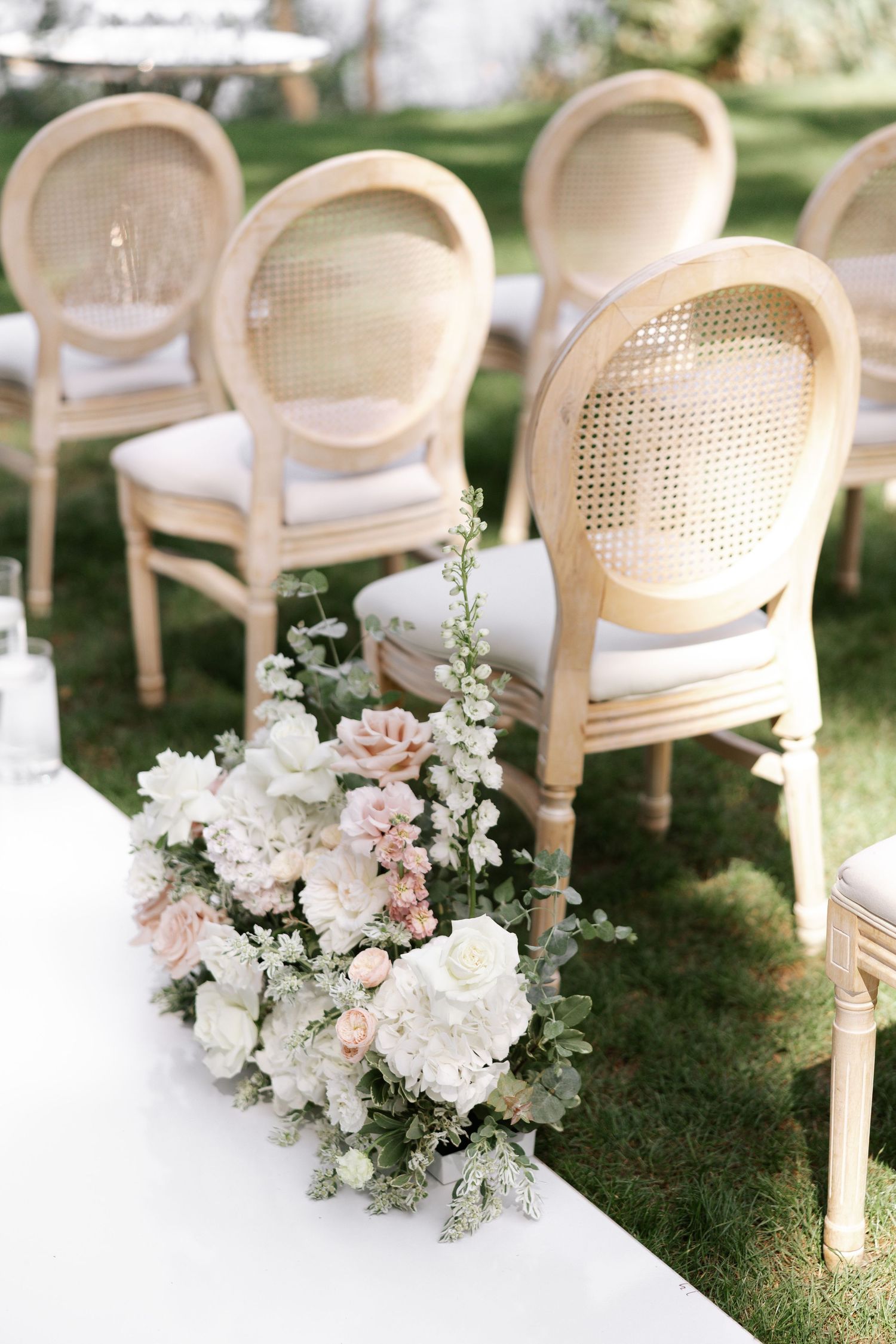 Vintage-style white cane chairs decorated with soft pink and white floral arrangements for outdoor ceremony.