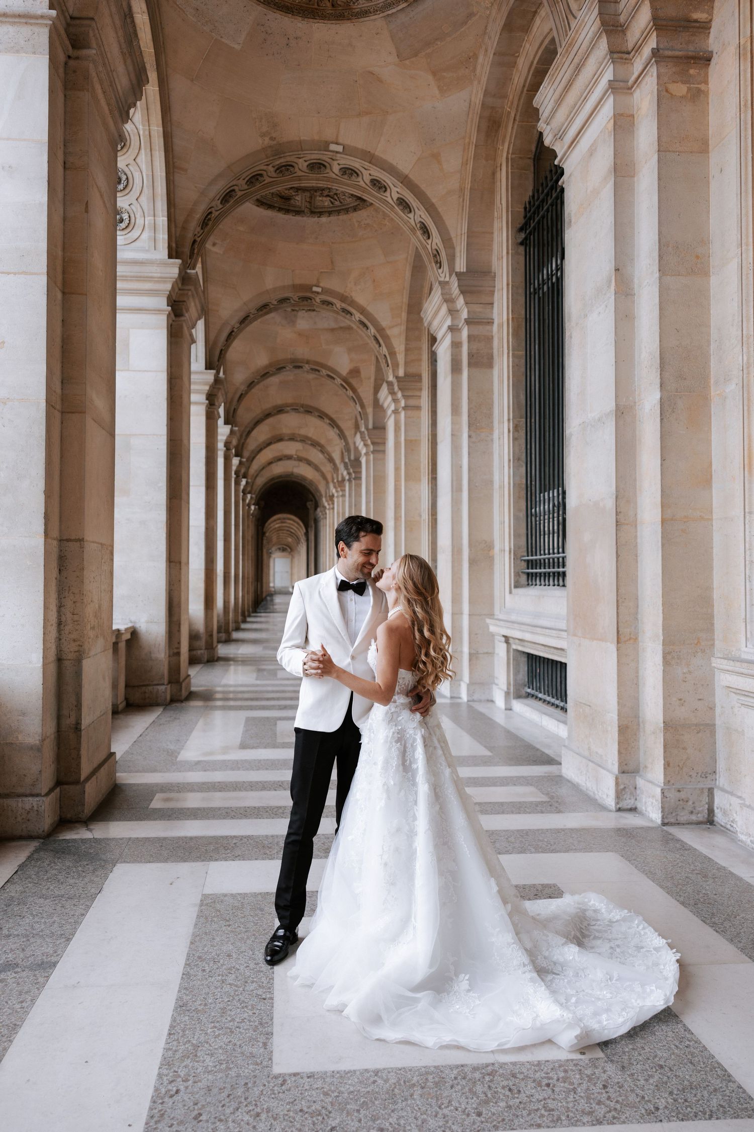 Wedding couple poses in ornate stone archway corridor in romantic European setting.