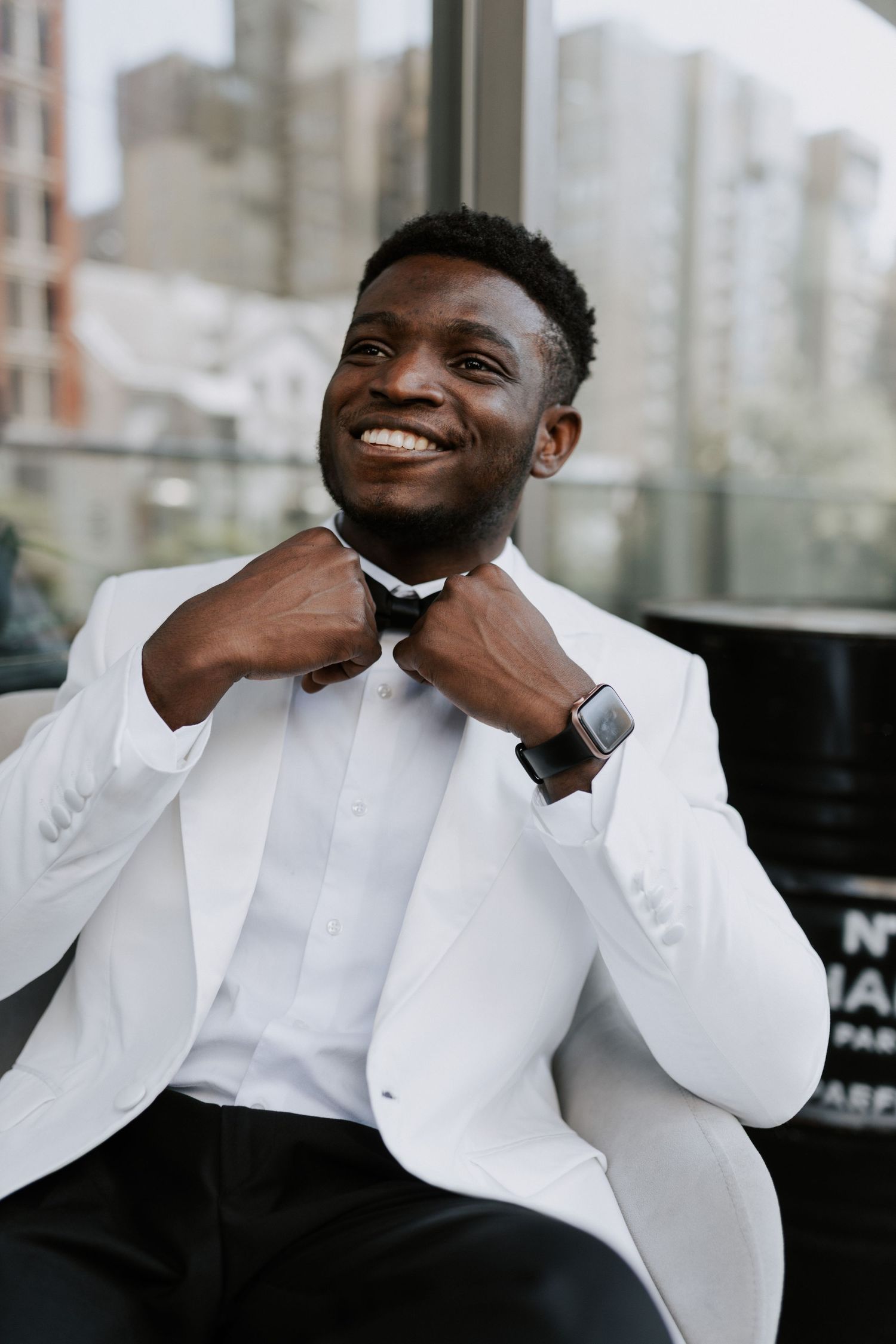 Groom adjusting bowtie while wearing crisp white tuxedo jacket against city backdrop.