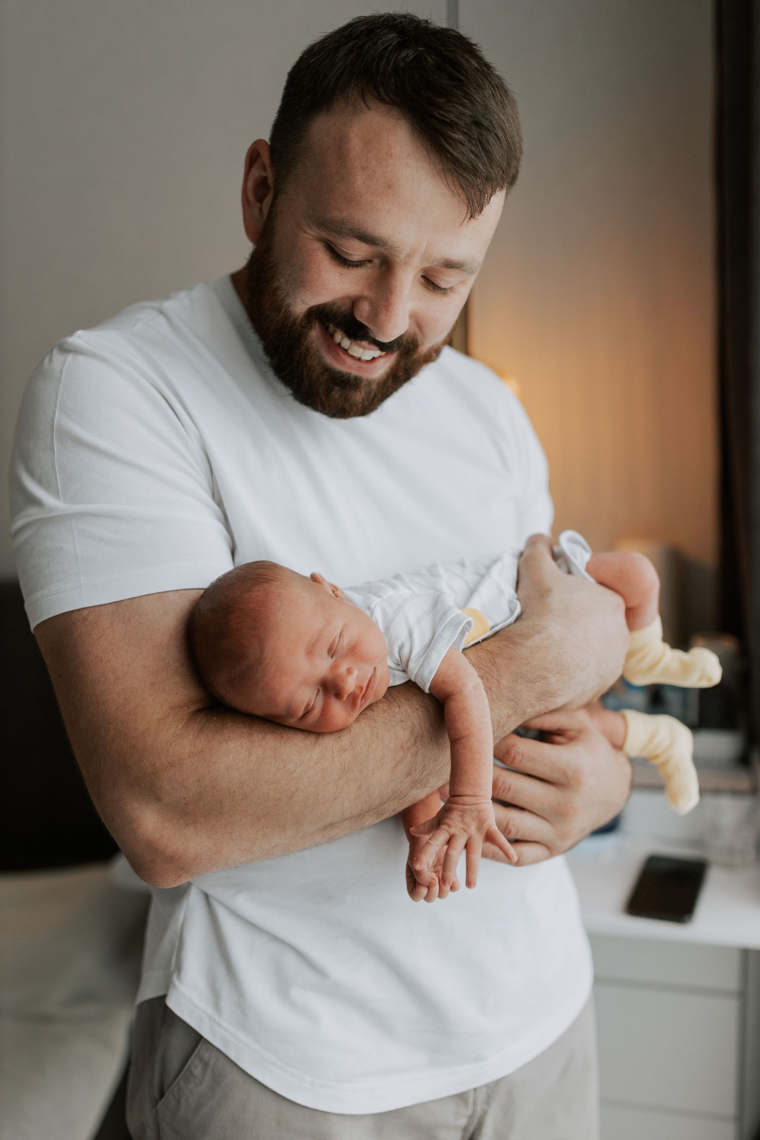 A bearded man in a white t-shirt tenderly holds a sleeping newborn baby with a small stuffed animal.