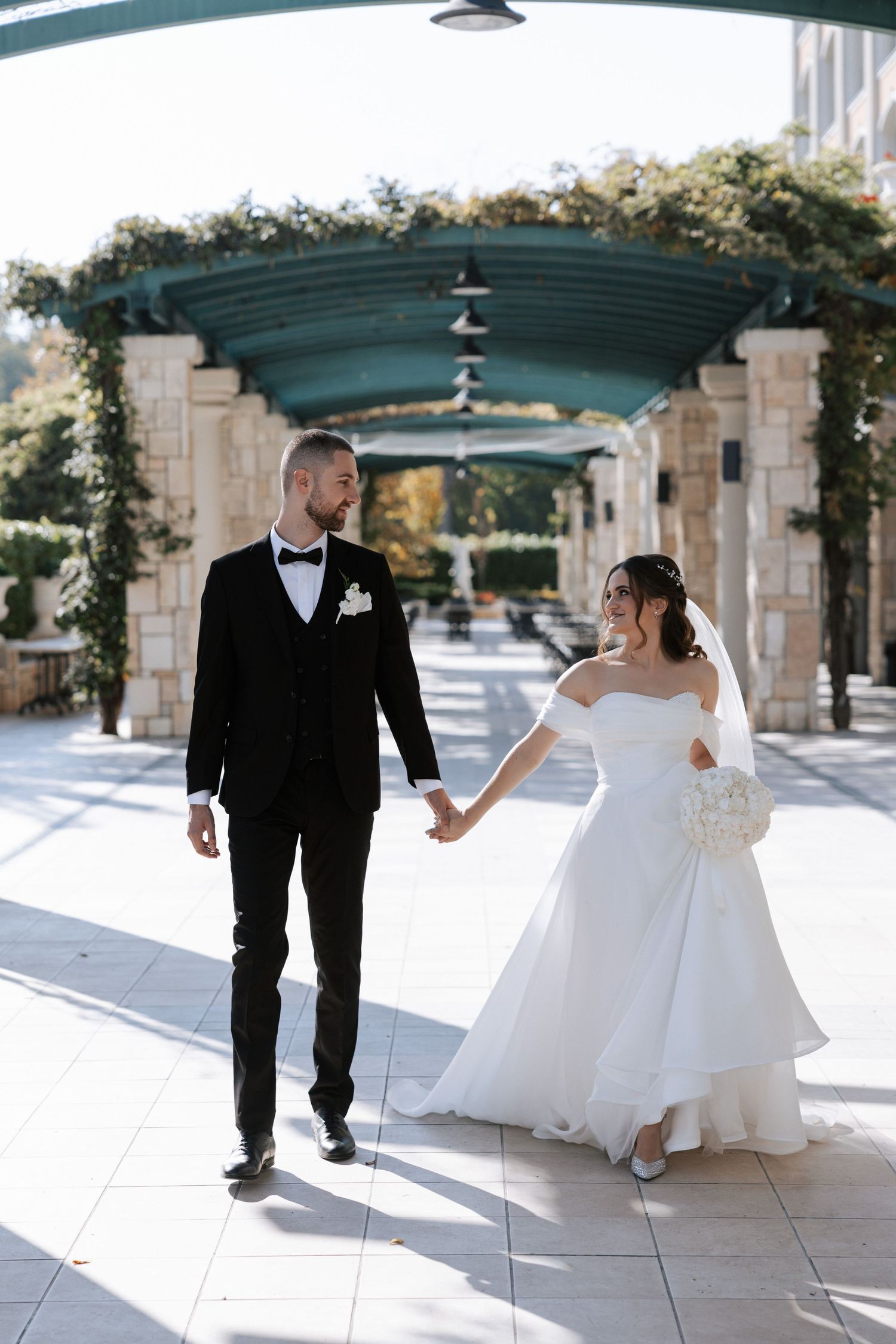 Couple walking hand in hand under elegant covered walkway decorated with greenery.