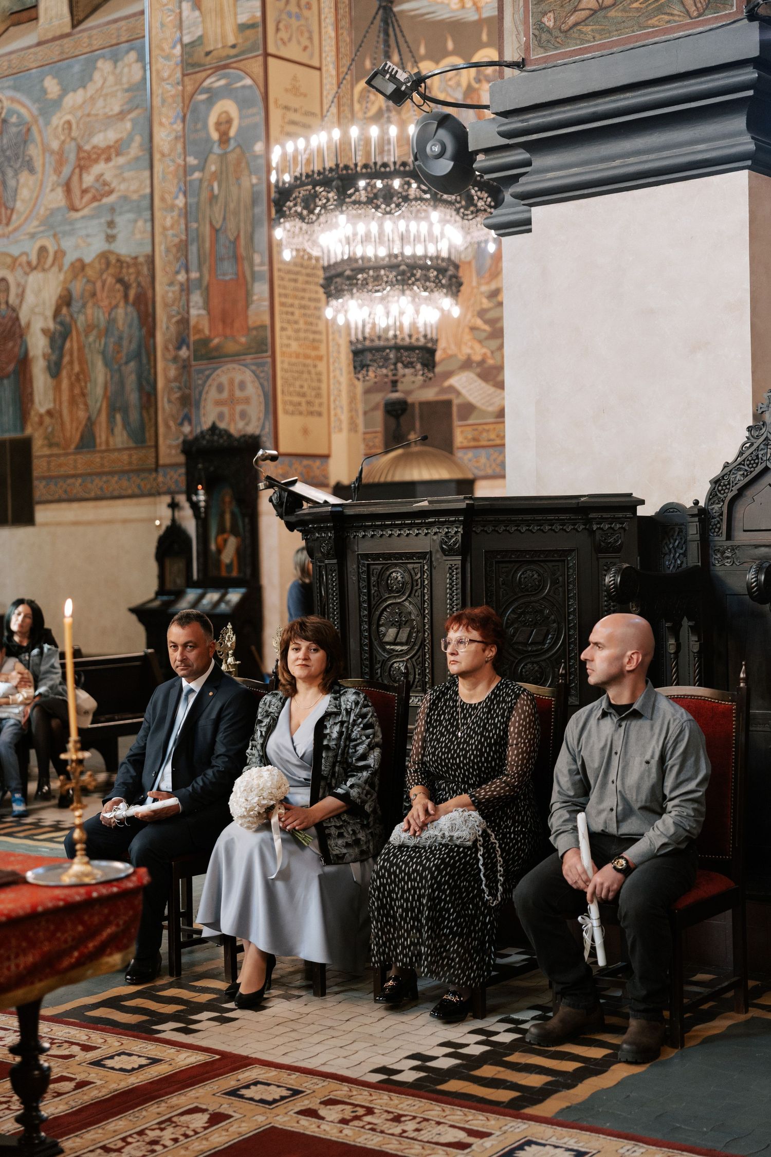 Wedding guests seated in ornate church pews with chandeliers and religious frescoes in background.