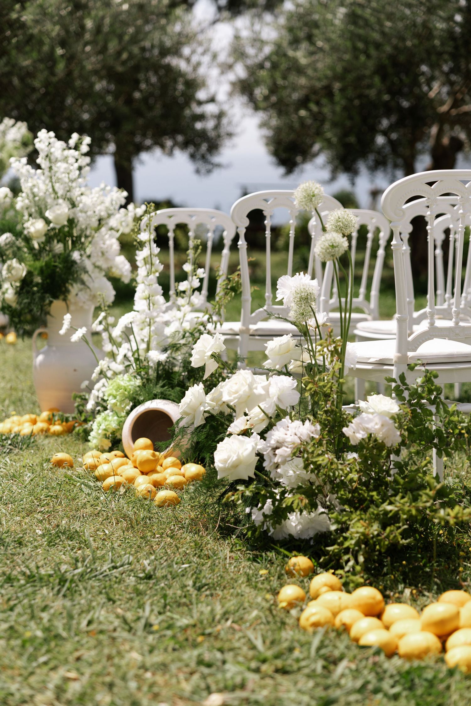 White wooden chairs line a garden path decorated with fresh oranges and white floral arrangements for an outdoor wedding ceremony.
