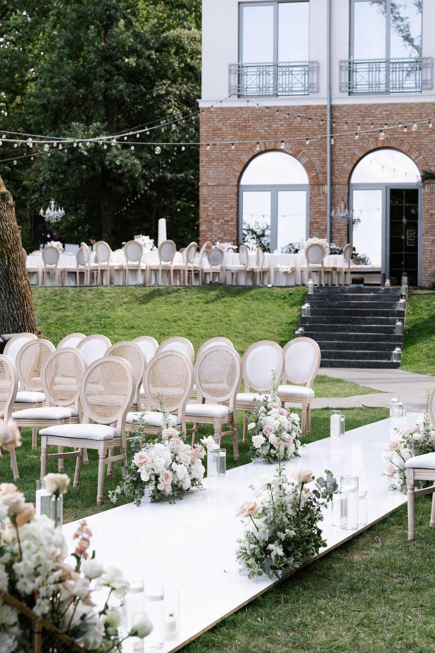 Elegant outdoor wedding ceremony setup with white chairs and floral arrangements along brick building.