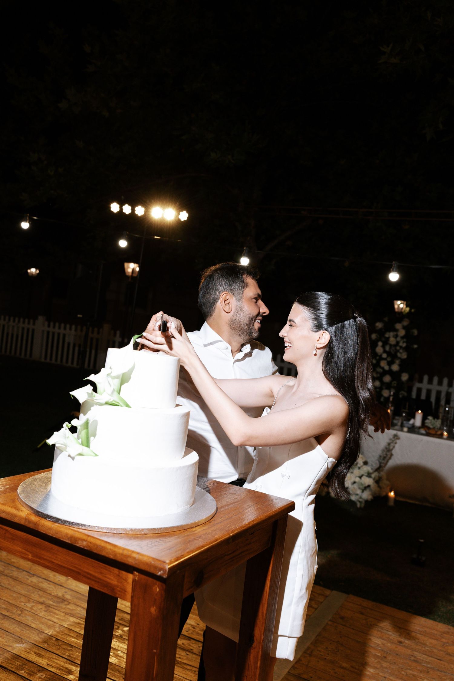 A couple shares a romantic moment while cutting their elegant white wedding cake at an outdoor evening reception.