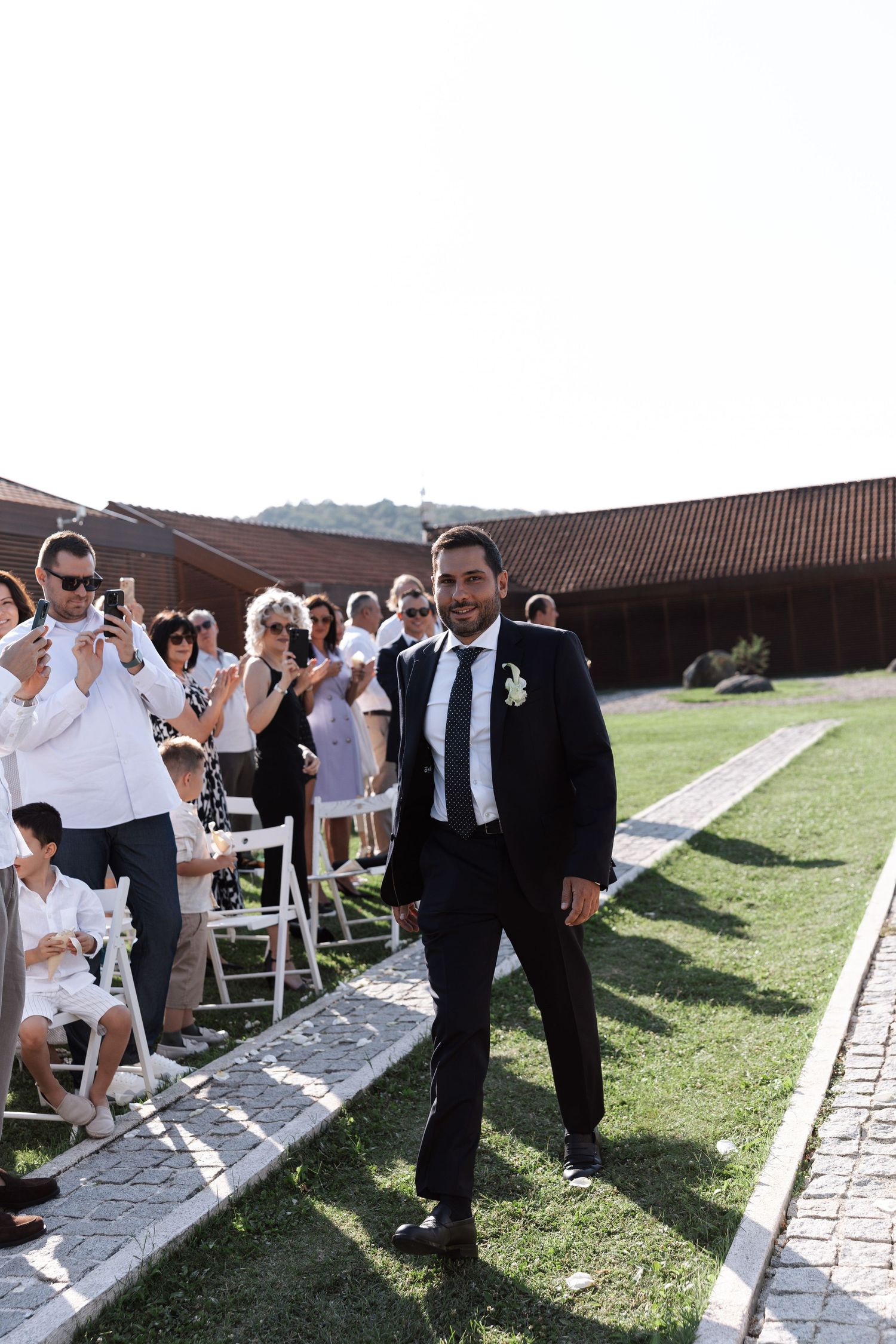 A groom in a black suit walks down an outdoor aisle while wedding guests in white clothing look on and take photos.