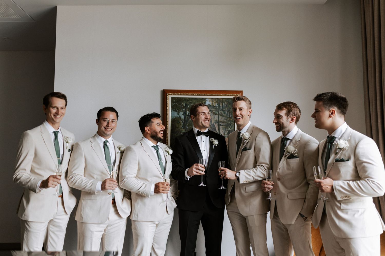 Wedding party in light suits posing with groom in black tuxedo indoors.