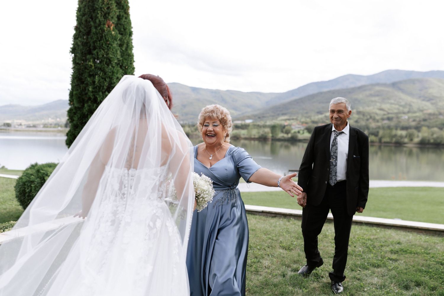 Joyful wedding moment captured near a lake with mountains and cypress trees in the background.