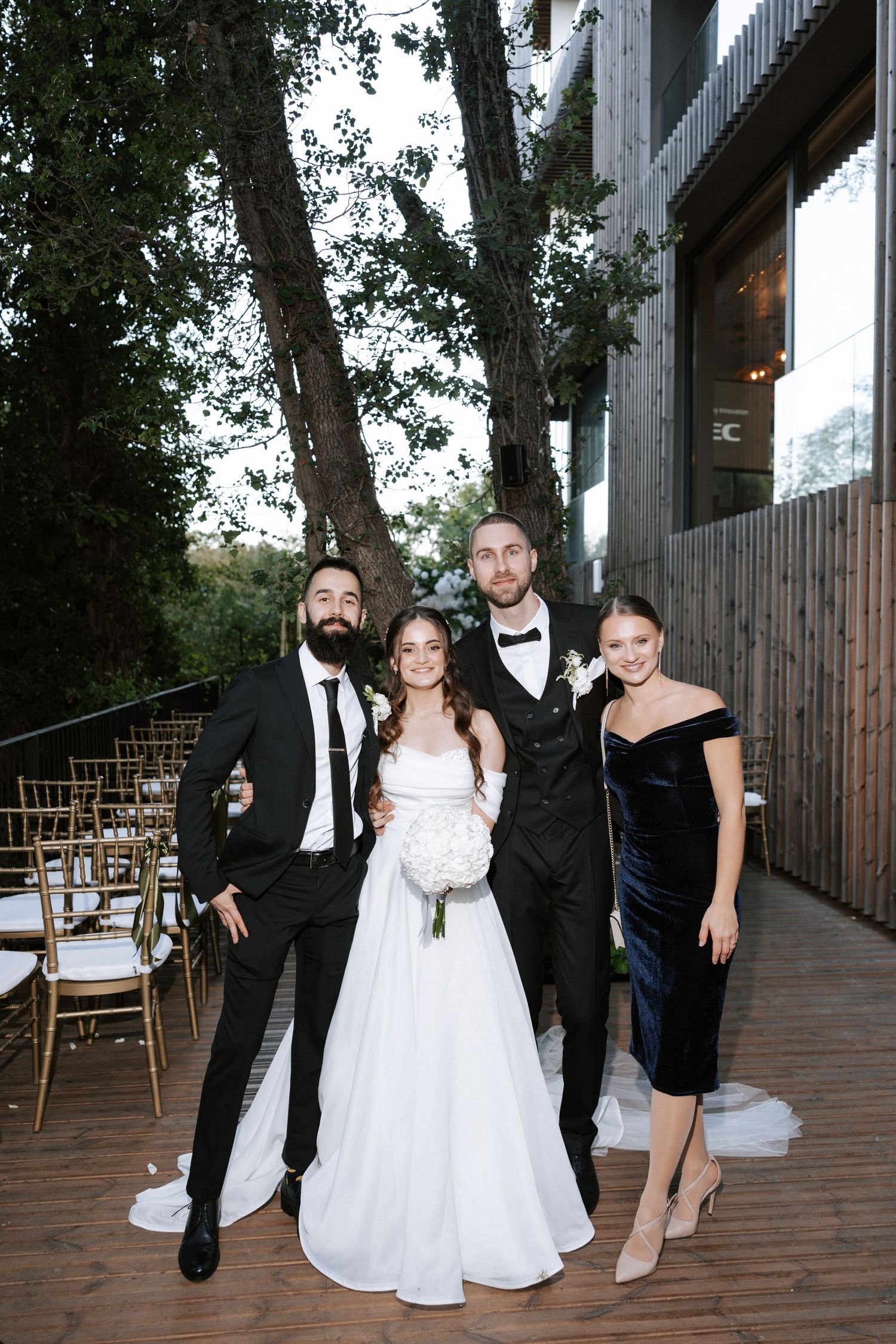 Formal wedding party poses on wooden deck with modern building backdrop.