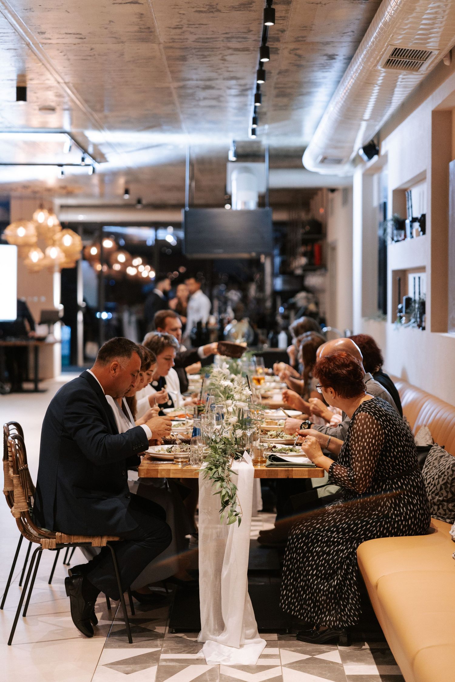 Wedding guests enjoying dinner at long tables in an industrial-style venue with modern lighting.