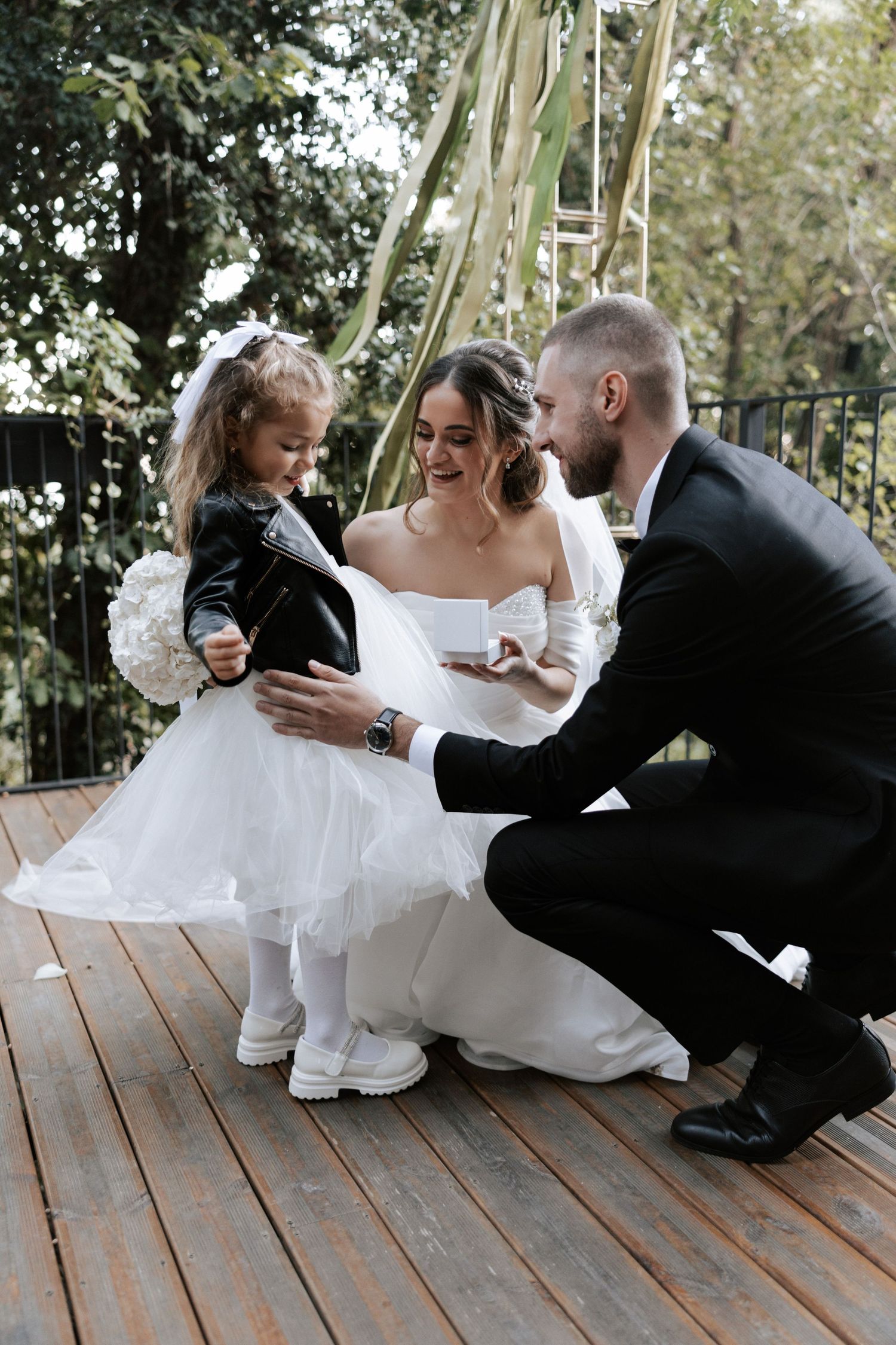 Wedding couple shares a tender moment with flower girl on wooden deck.