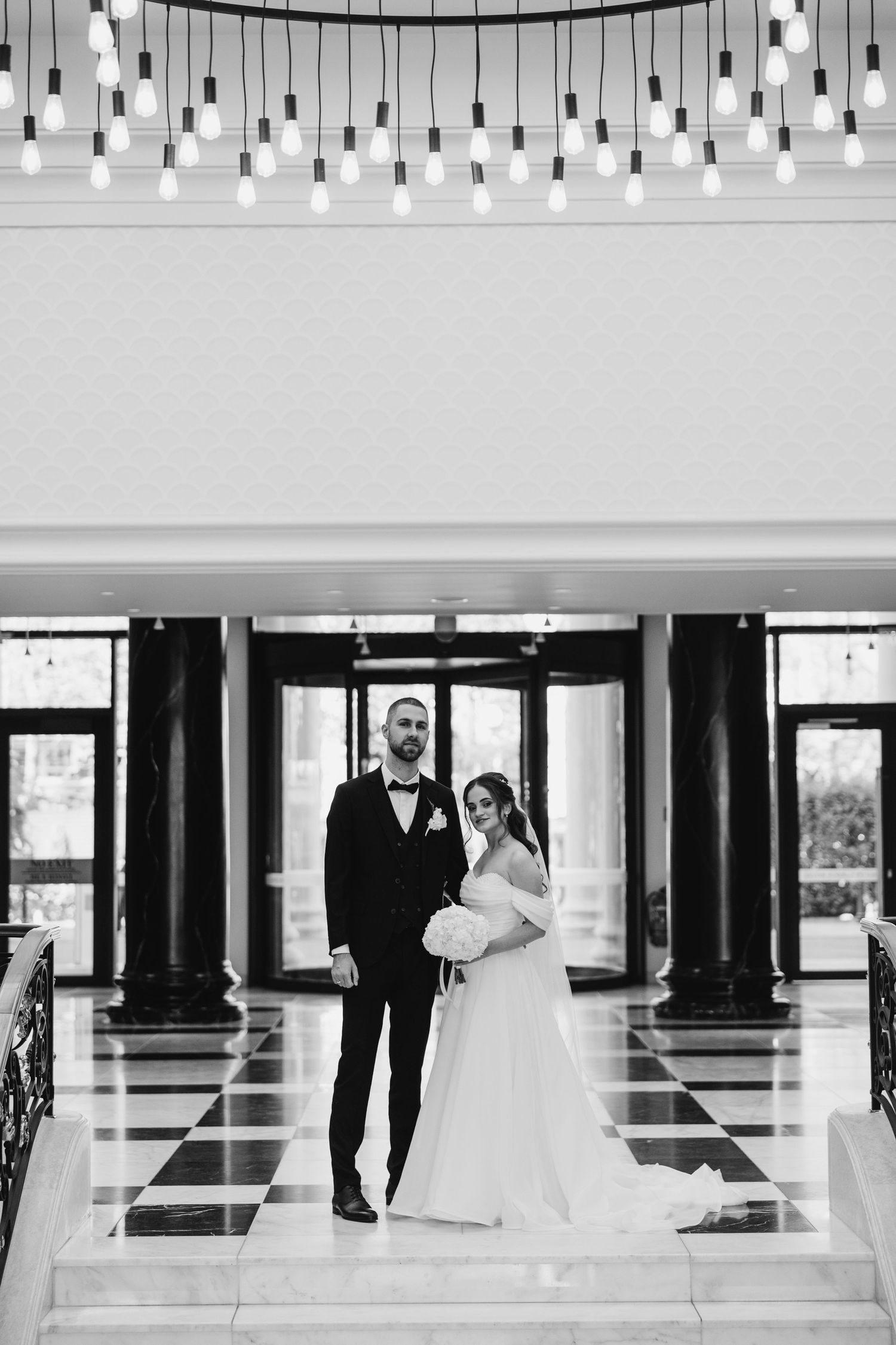 Wedding couple stands under pendant lights on checkered floor in elegant venue entrance.