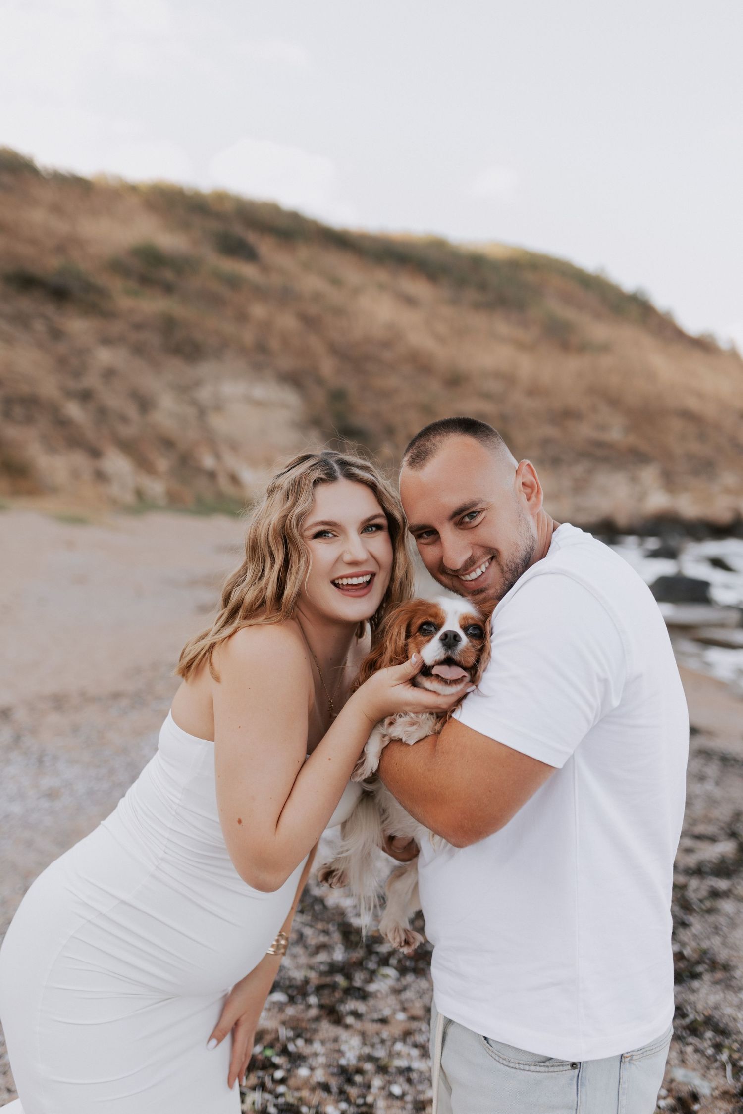 A couple poses with their small dog on a rocky beach while wearing white outfits.