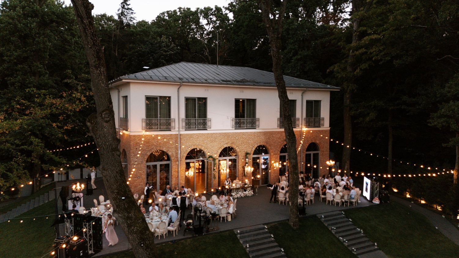 Evening wedding reception at illuminated brick villa with string lights and guests dining outdoors.