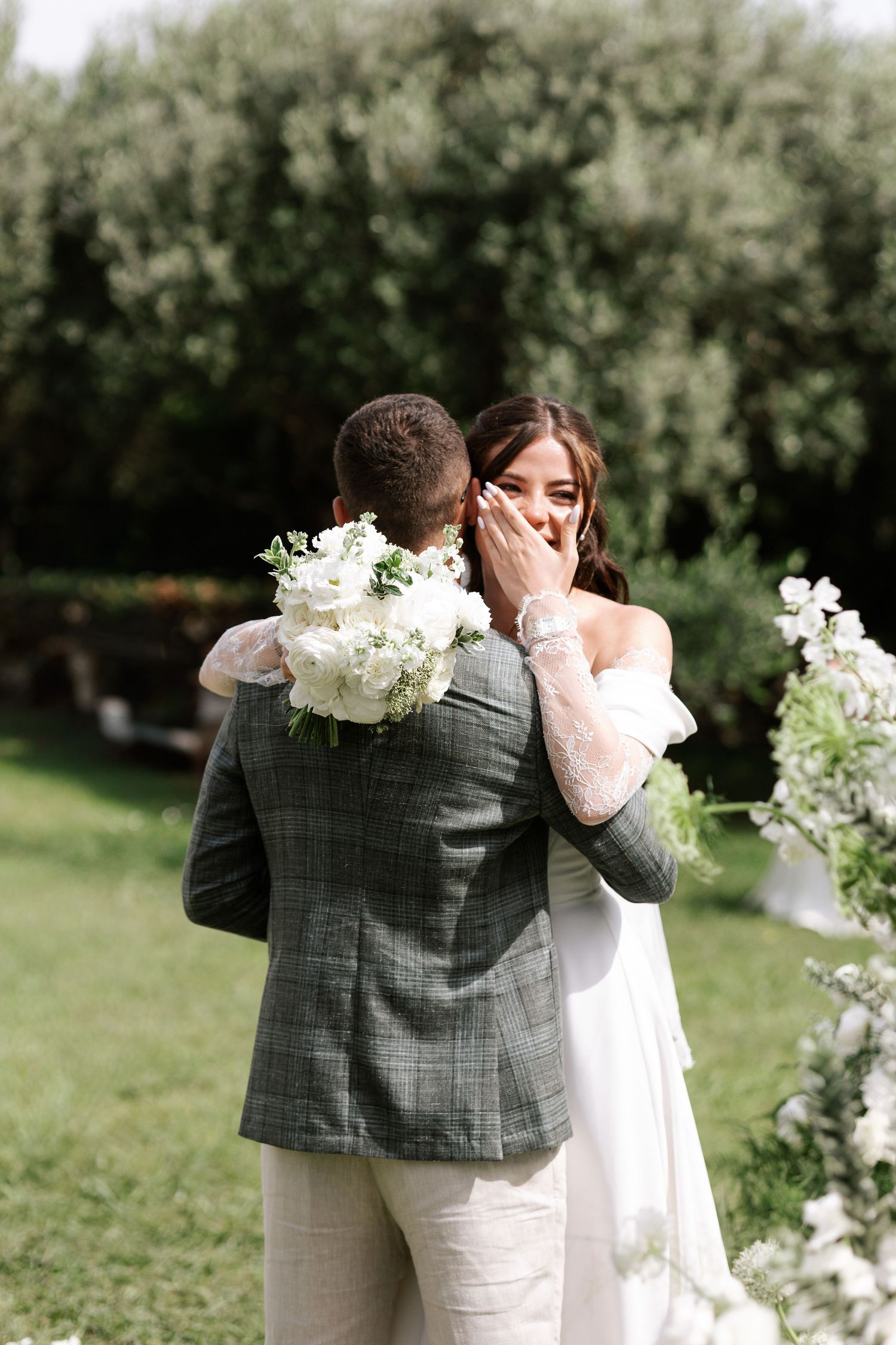 A couple embraces during their first look moment in a lush garden setting with white floral decorations.