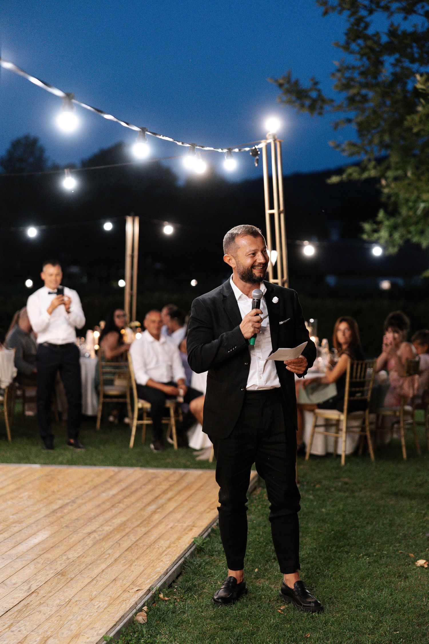String lights illuminate an outdoor evening wedding reception with a wooden dance floor set up on grass.