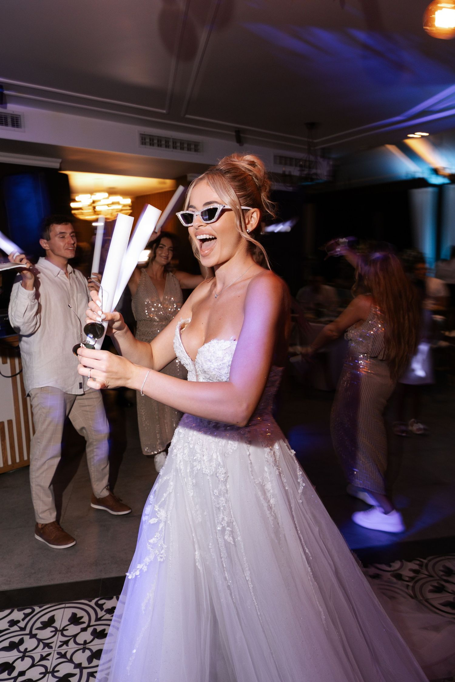 A bride in a white tulle gown and sunglasses dances at her wedding reception holding light-up glow sticks.