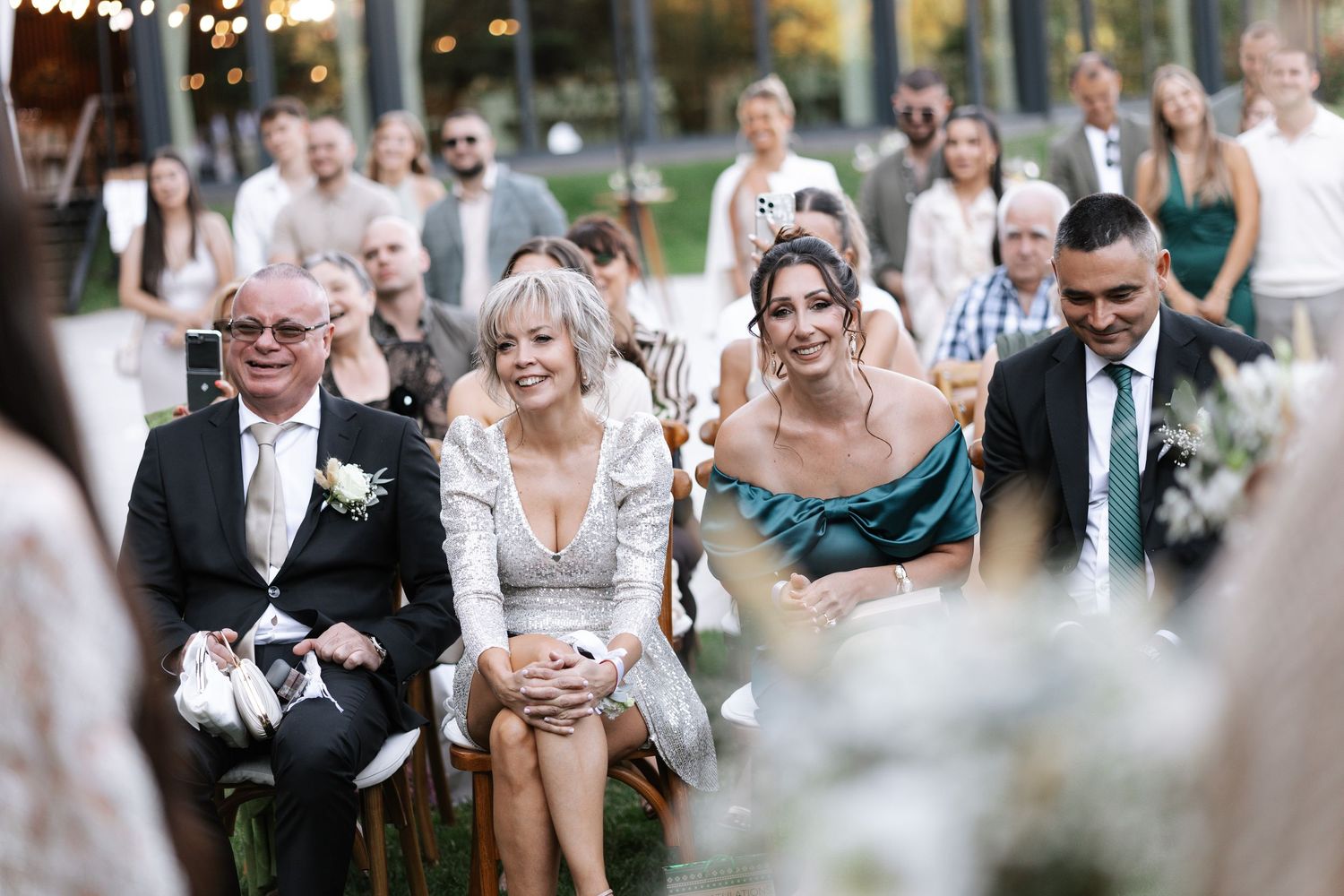 Wedding guests laugh and react emotionally during an outdoor ceremony at a garden venue.