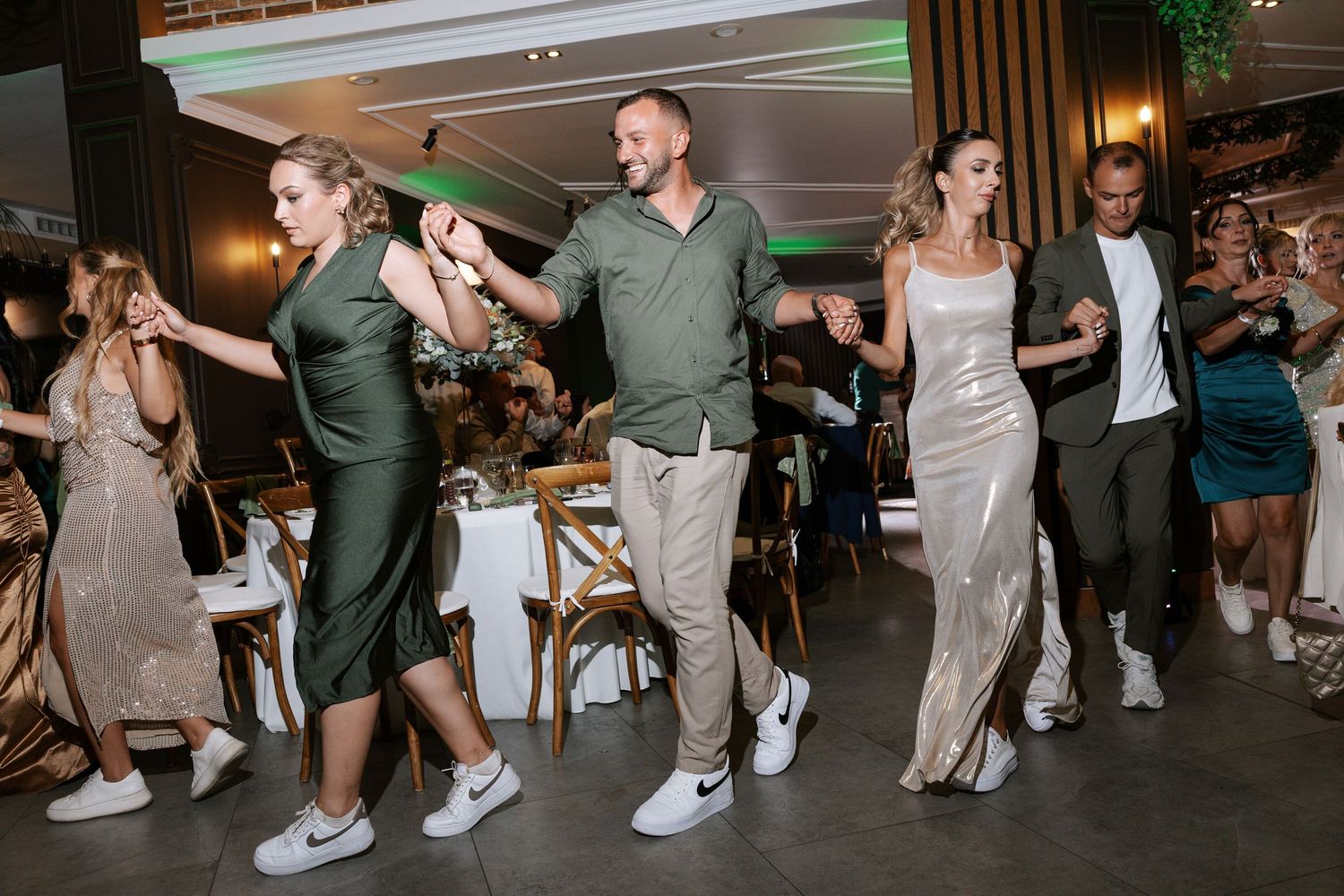 Wedding guests dance in a line wearing coordinated green and neutral outfits at an indoor reception venue.