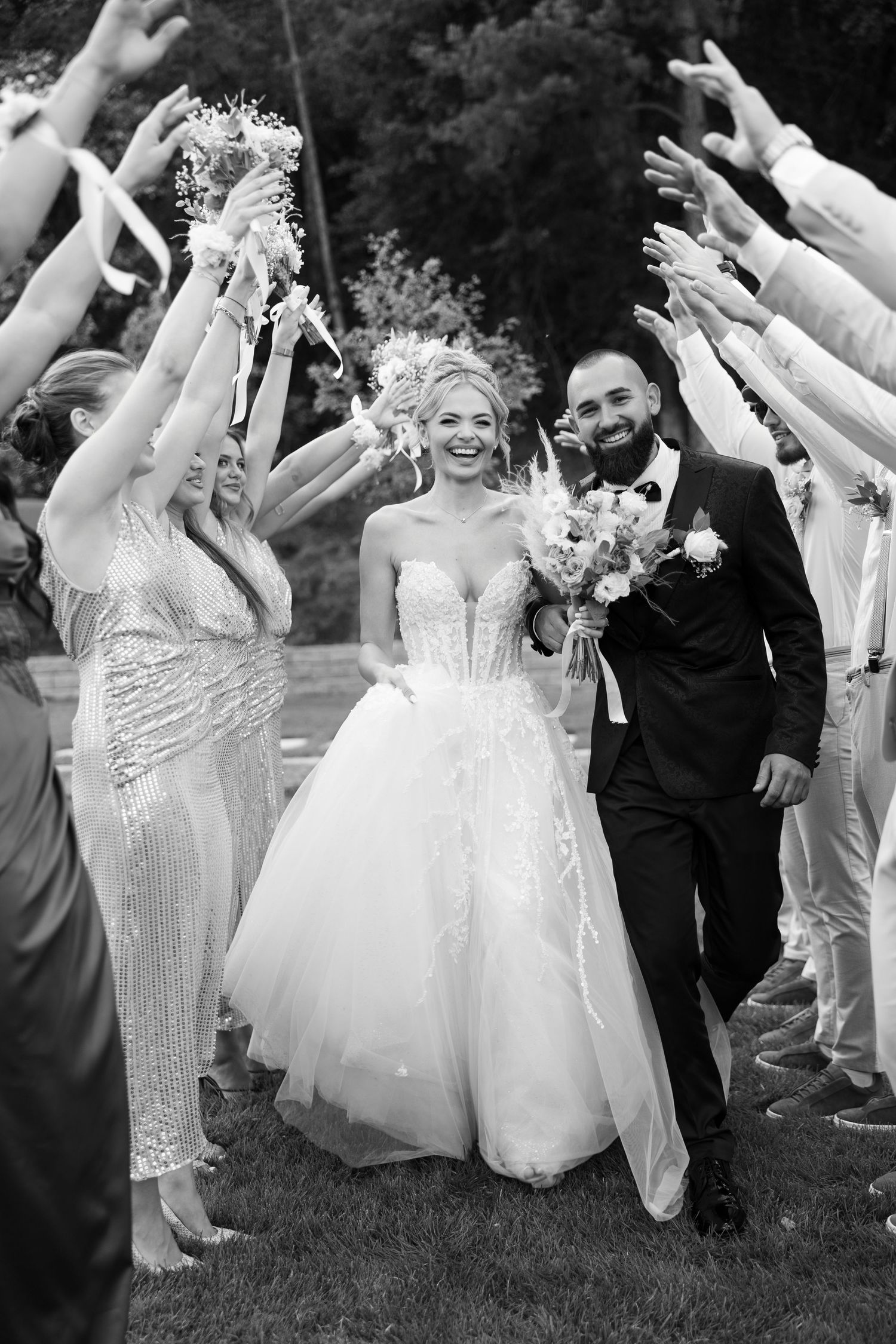 Wedding party celebrates joyfully on grass with raised arms and flowing white dresses in black and white photo.