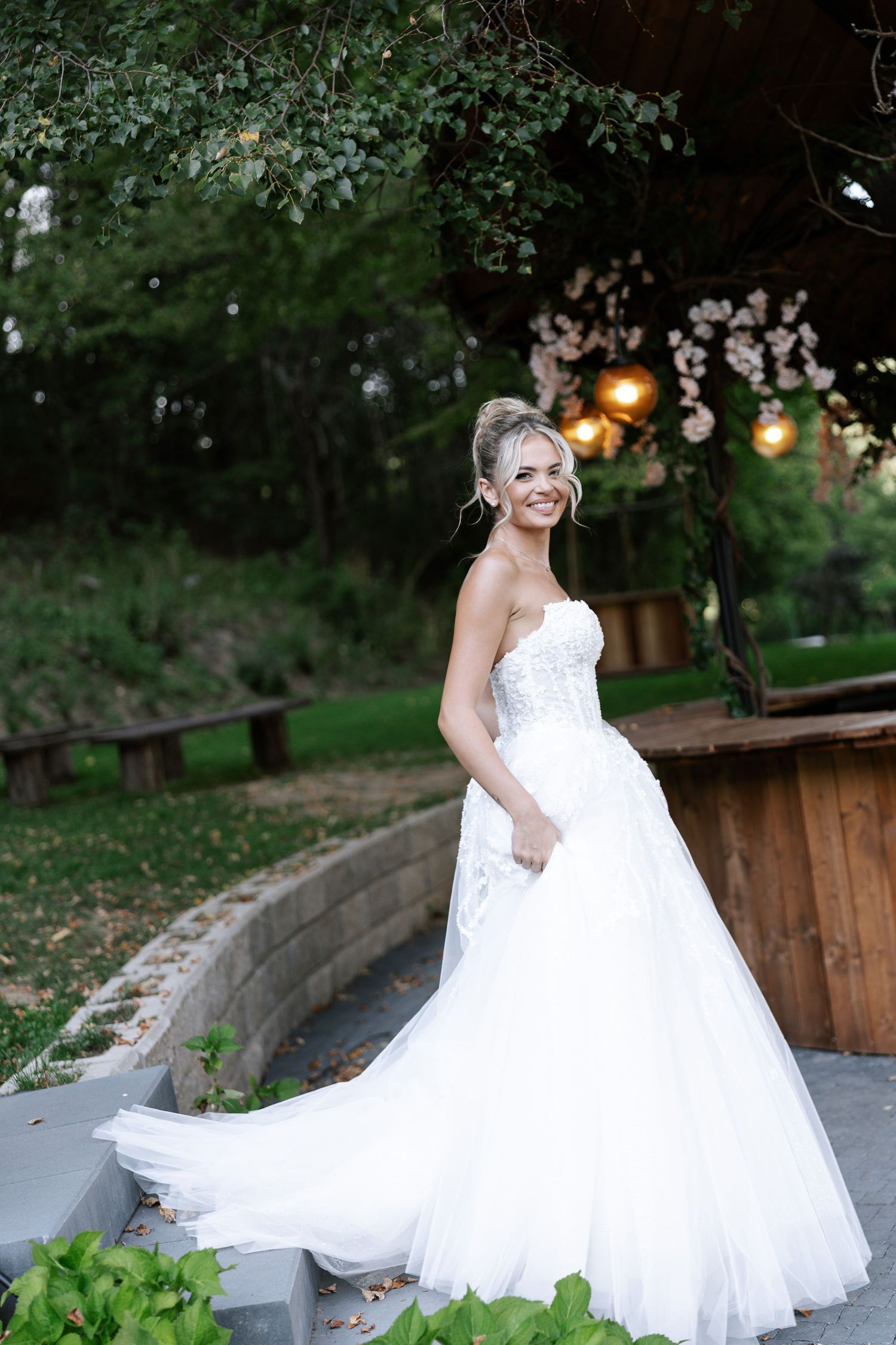 A bride in a flowing white ball gown poses gracefully in a garden setting with string lights and greenery in the background.