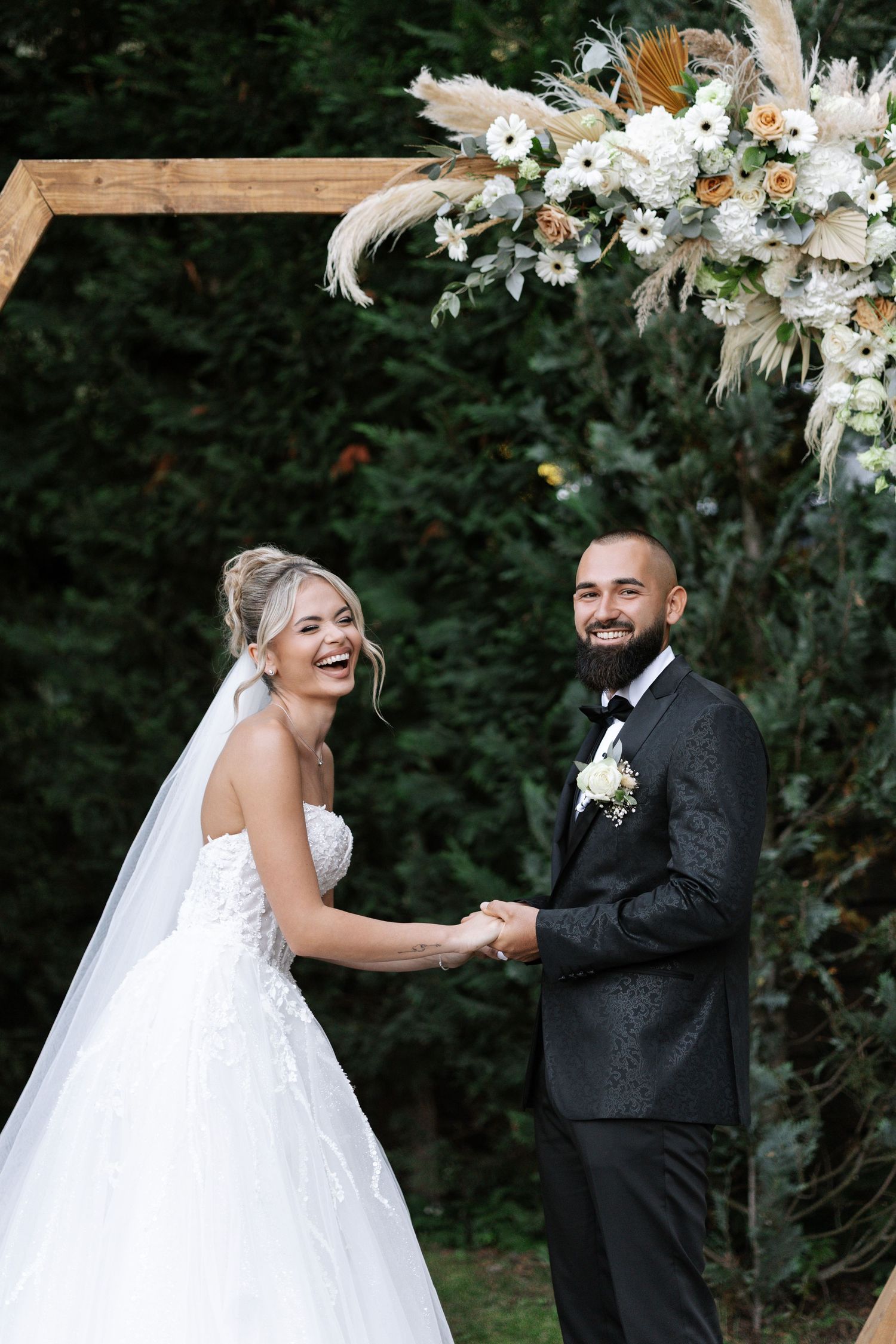 A happy couple shares a joyful moment during their outdoor wedding ceremony under floral decorations.
